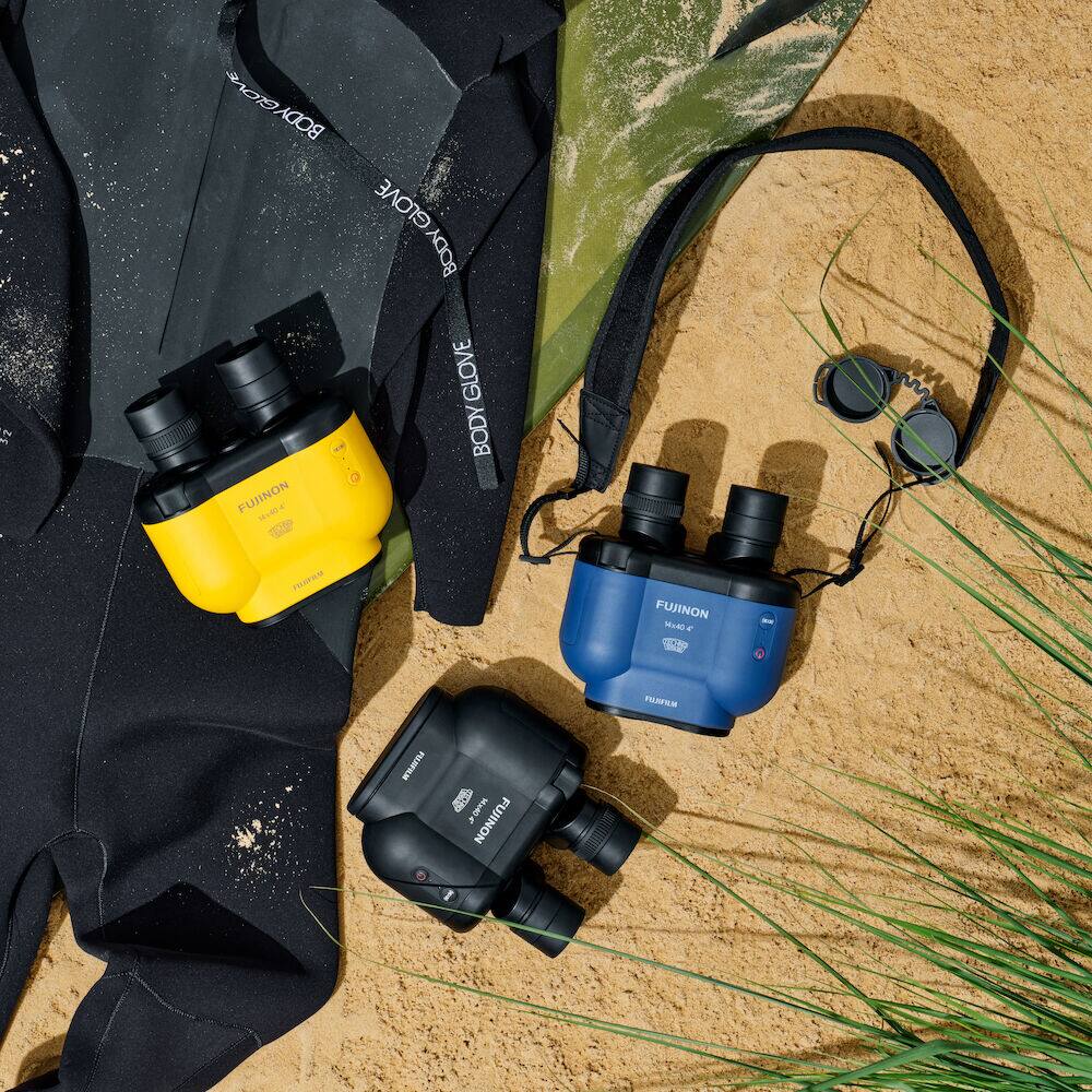 The image features a variety of items, including a yellow and black camera, a blue camera, and a black wetsuit. There is also a pair of sunglasses and a body glove present in the scene. The items are placed on a sandy surface, possibly a beach.