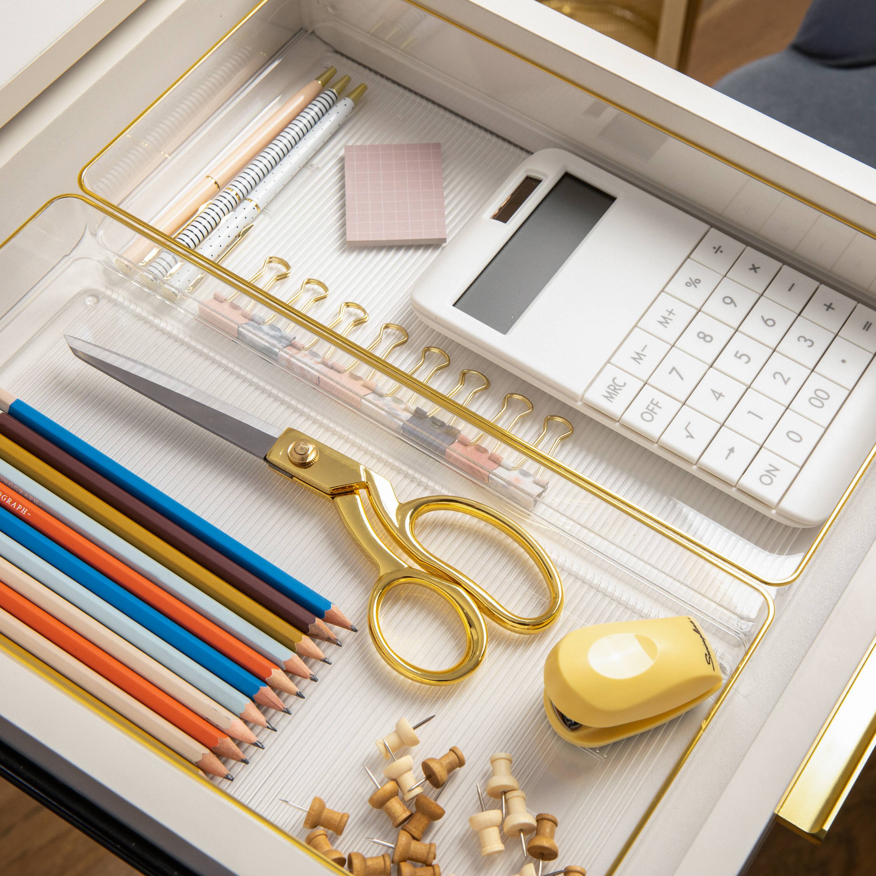 The image shows a white desk with a drawer open, revealing various office supplies. Inside the drawer, there are multiple pencils, a pair of scissors, a calculator, and a cell phone. The pencils are arranged in different positions, with some lying horizontally and others vertically. The scissors are placed near the center of the drawer, while the calculator and cell phone are located towards the right side of the drawer. The assortment of items in the drawer indicates that it is well-stocked for various office tasks and projects.