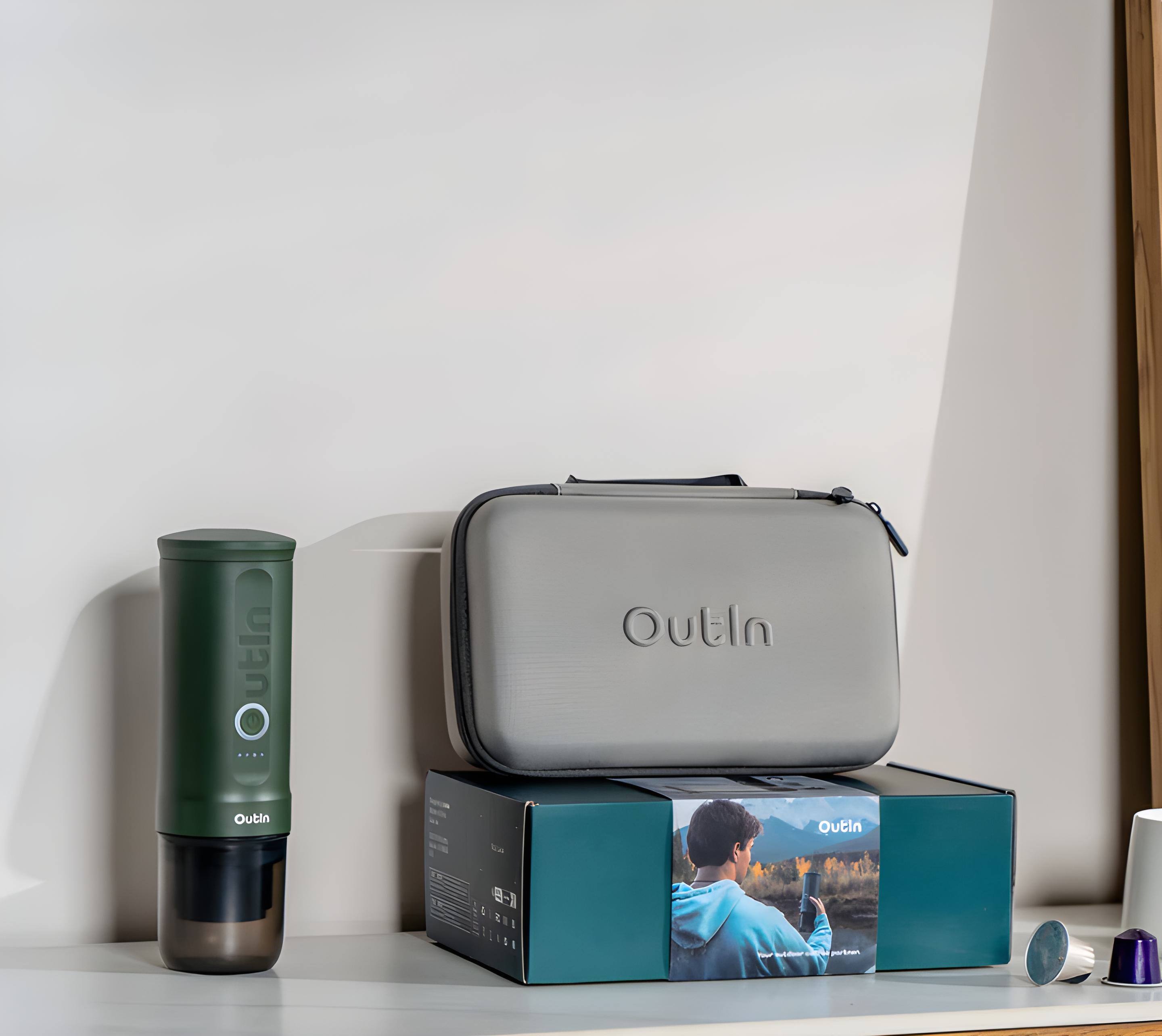 Outliner is a product that is displayed on a shelf. It is a small, grey case that is placed next to a green object, which appears to be a coffee mug. The Outliner case is sitting on a table, and it is accompanied by a box that contains a picture of a man.