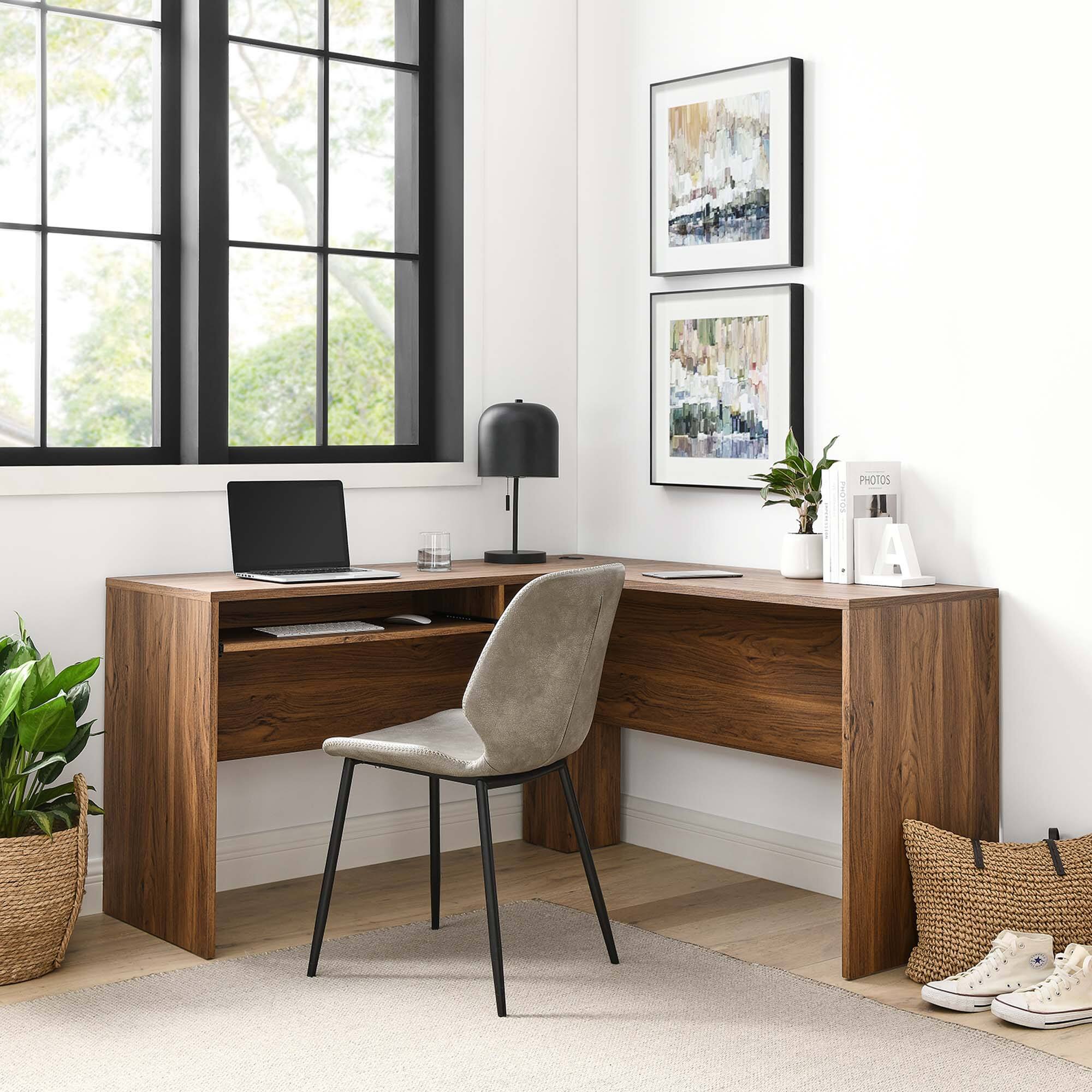 A home office setup featuring a wooden desk with a laptop, a black desk lamp, and a small plant. On the desk, there are two framed abstract paintings and a white organizer labeled "PHOTOS." A gray chair is positioned in front of the desk, and a pair of white sneakers is placed on the floor beside a woven basket. The room has a large window with a view of greenery outside.