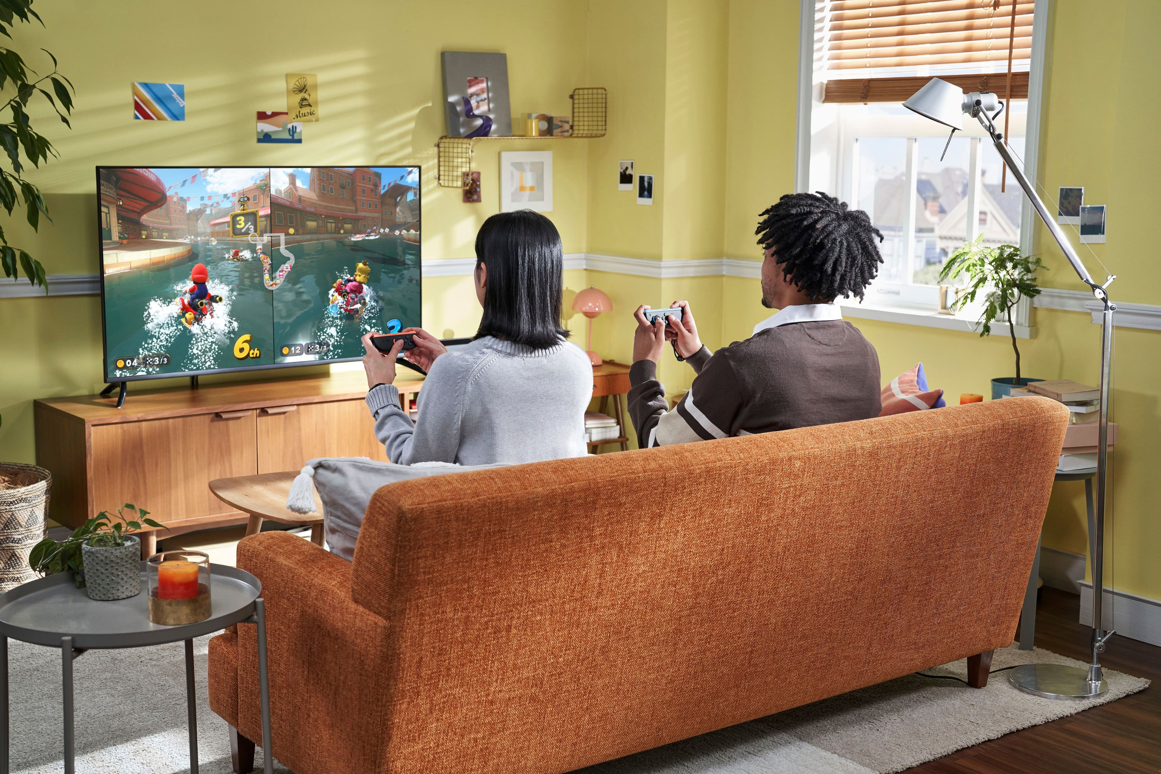 The image shows a man and a woman sitting on a couch, playing a video game on a TV. They are both holding game controllers, and the TV is located on the left side of the room. The couch is situated in the middle of the room, and there are two potted plants nearby, one on the left side and the other on the right side. The scene appears to be a cozy and comfortable living space where the couple can enjoy their gaming session.