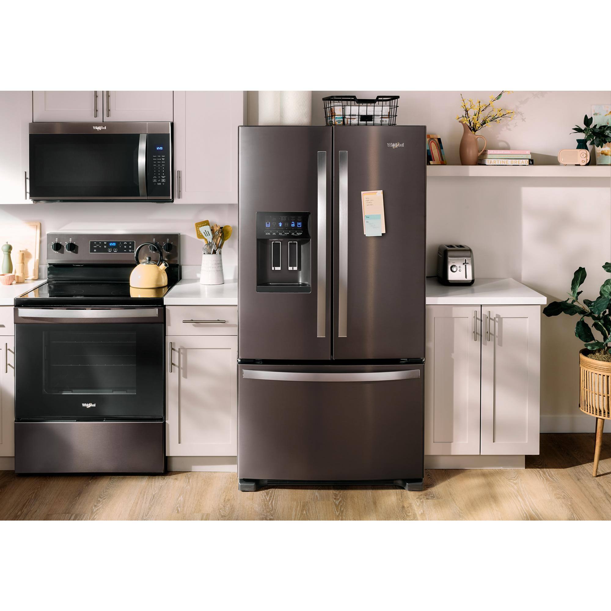 The image shows a kitchen with a metallic refrigerator, a stove, and a microwave. The refrigerator is a large, two-door model with a water dispenser and ice maker. The stove is a black, electric model with a toaster oven on top of it. The microwave is positioned above the stove. The kitchen also features a sink and a potted plant.