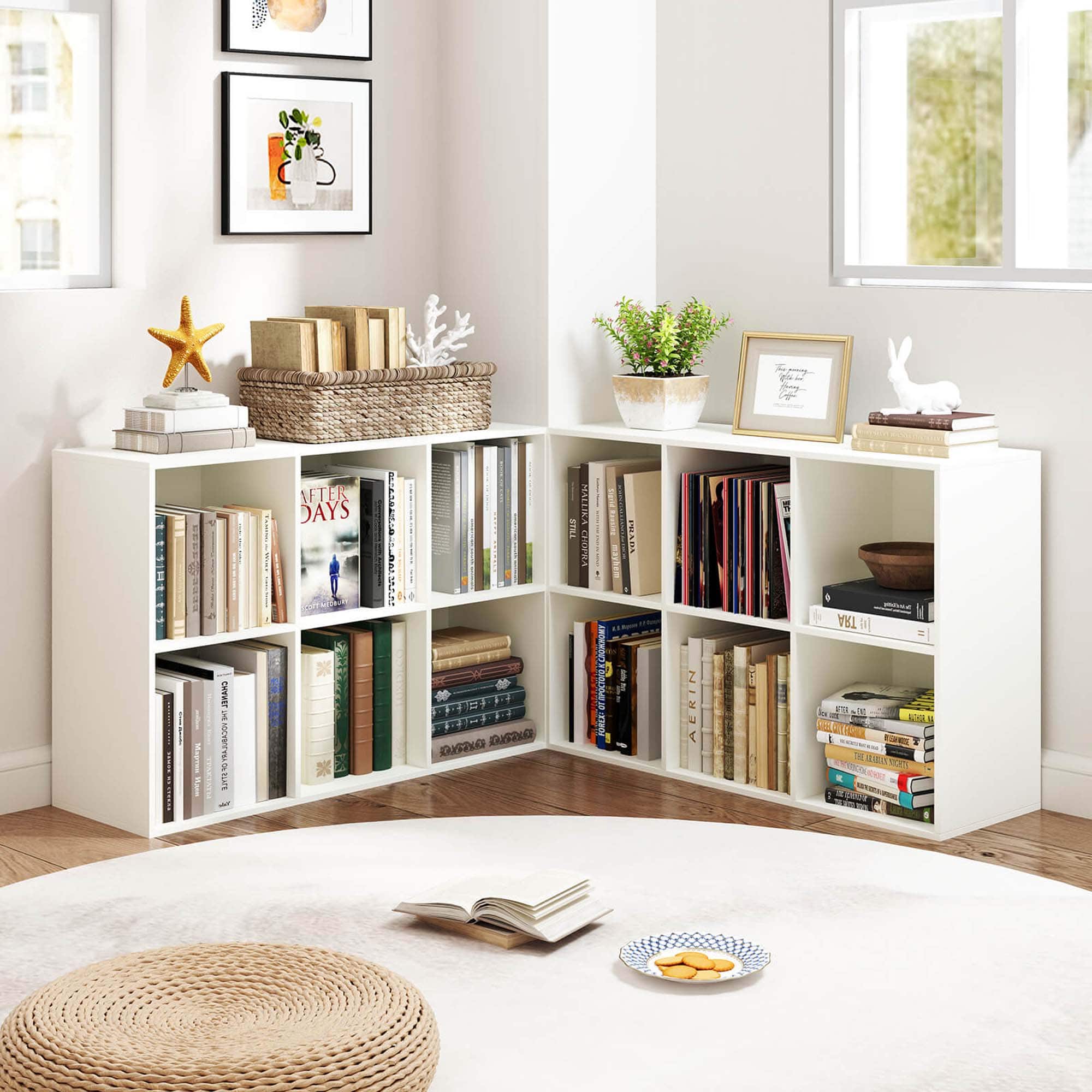The image features a cozy living room with a white bookshelf filled with books and a wicker basket. There are several books on the bookshelf, including one titled "After Days." A small table is also present in the room, with a bowl and a plate on it. Additionally, there are two vases in the room, one placed on the bookshelf and the other on the table. A potted plant is situated near the bookshelf, adding a touch of greenery to the space.