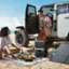 The image shows a man and a woman sitting on a blanket on the beach, with a truck parked nearby. The man is holding a bottle, and there are several other bottles and a cup scattered around them. They seem to be enjoying their time together, possibly sharing a meal or drinks. The scene conveys a sense of relaxation and leisure, as the couple spends time together in a beautiful outdoor setting.
