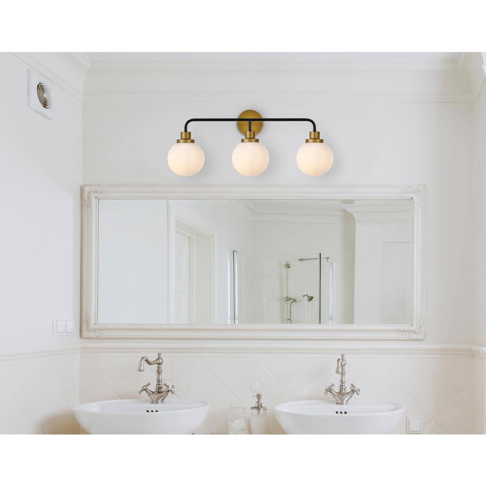 The image shows a bathroom with a large mirror above two white sinks. The mirror has a classic frame, and there is a three-bulb light fixture with a black and gold design above it. The faucets are silver with a traditional design. The walls are white, and there is a shower visible in the reflection of the mirror.