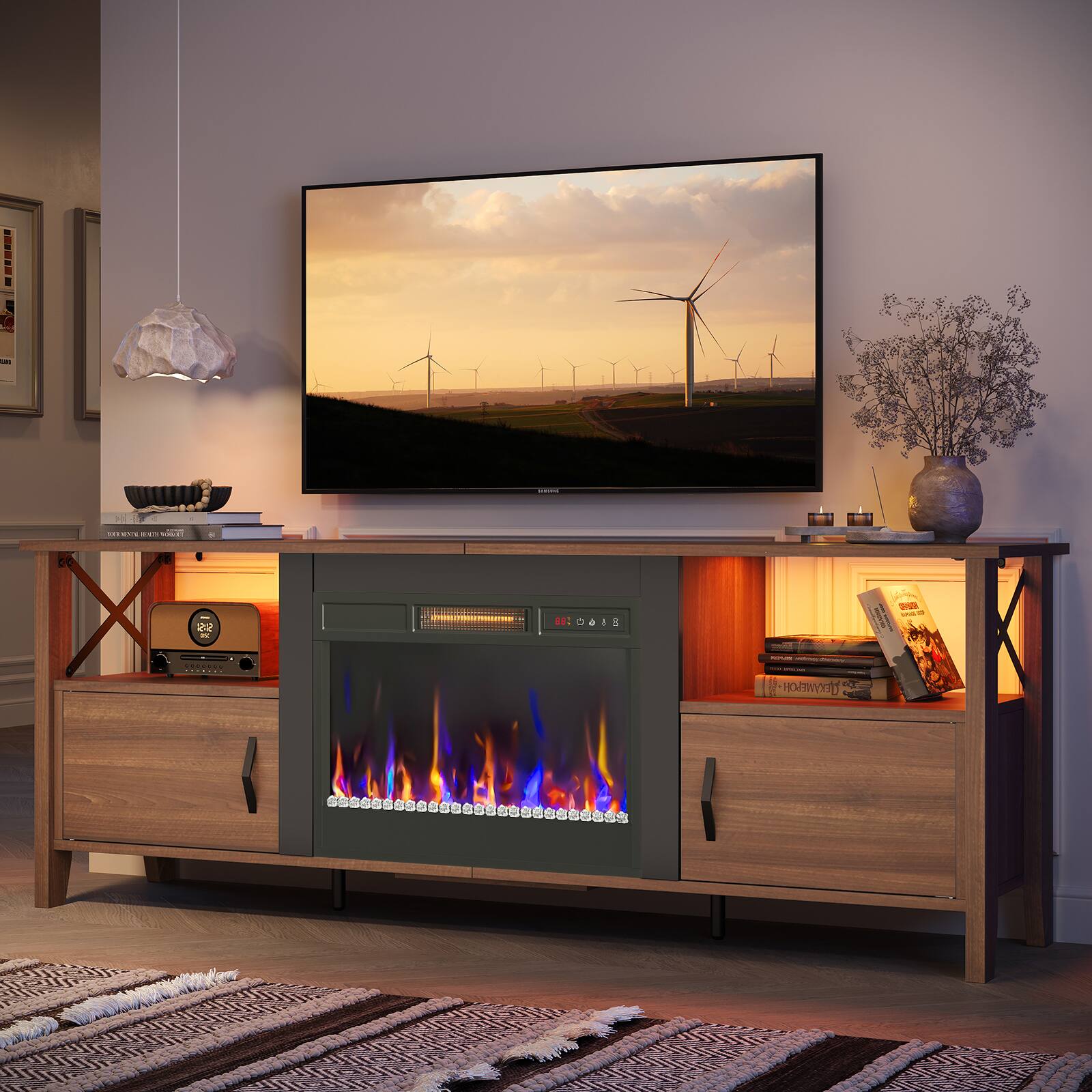 The image features a living room with a flat screen TV mounted on the wall. The TV is displaying a picture of windmills. The room also has a fireplace, which is lit, adding warmth and ambiance to the space. The room is furnished with a couch, a chair, and a potted plant. There are several books scattered around the room, and a vase is placed on a surface. The overall atmosphere of the living room is cozy and inviting.