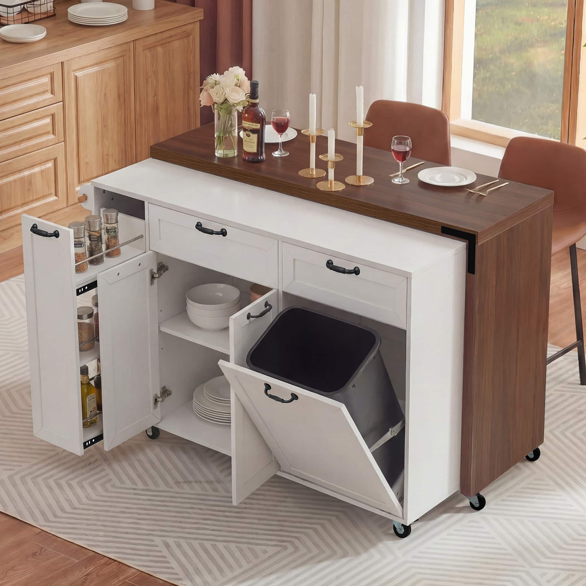The image shows a modern kitchen cart with a wooden top and white drawers. The cart has multiple compartments, including a pull-out drawer with a trash bin, shelves with plates and bowls, and a spice rack. On the top, there are candles, a bottle of wine, a glass of red wine, and a vase with flowers. The cart is on wheels and is positioned near a dining table with chairs.