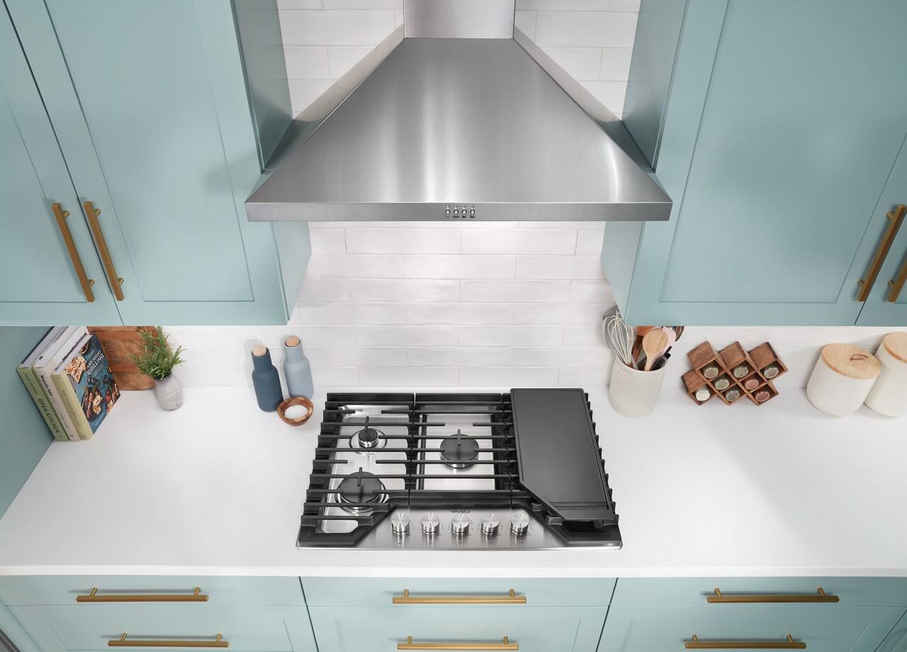 The image features a clean and organized kitchen with a white countertop and a stove top oven. The oven is positioned under a stainless steel ventilation hood, which is an essential feature for any kitchen. The countertop is adorned with various items, including a collection of books, a few bottles, and a potted plant. Additionally, there are several knives placed on the countertop, and a vase is also visible. The kitchen appears to be well-maintained and ready for use.
