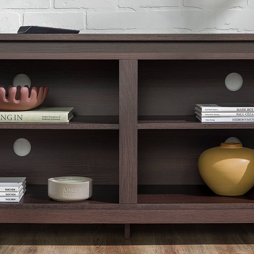 The image features a wooden bookshelf with various items on it. There are several books placed on different shelves, including a book titled "Mark Rothko" and another titled "Paul Cezanne." Additionally, there is a bowl on the shelf, and a vase is placed on the bottom shelf. The bookshelf is situated in front of a brick wall, giving the scene a cozy and inviting atmosphere.