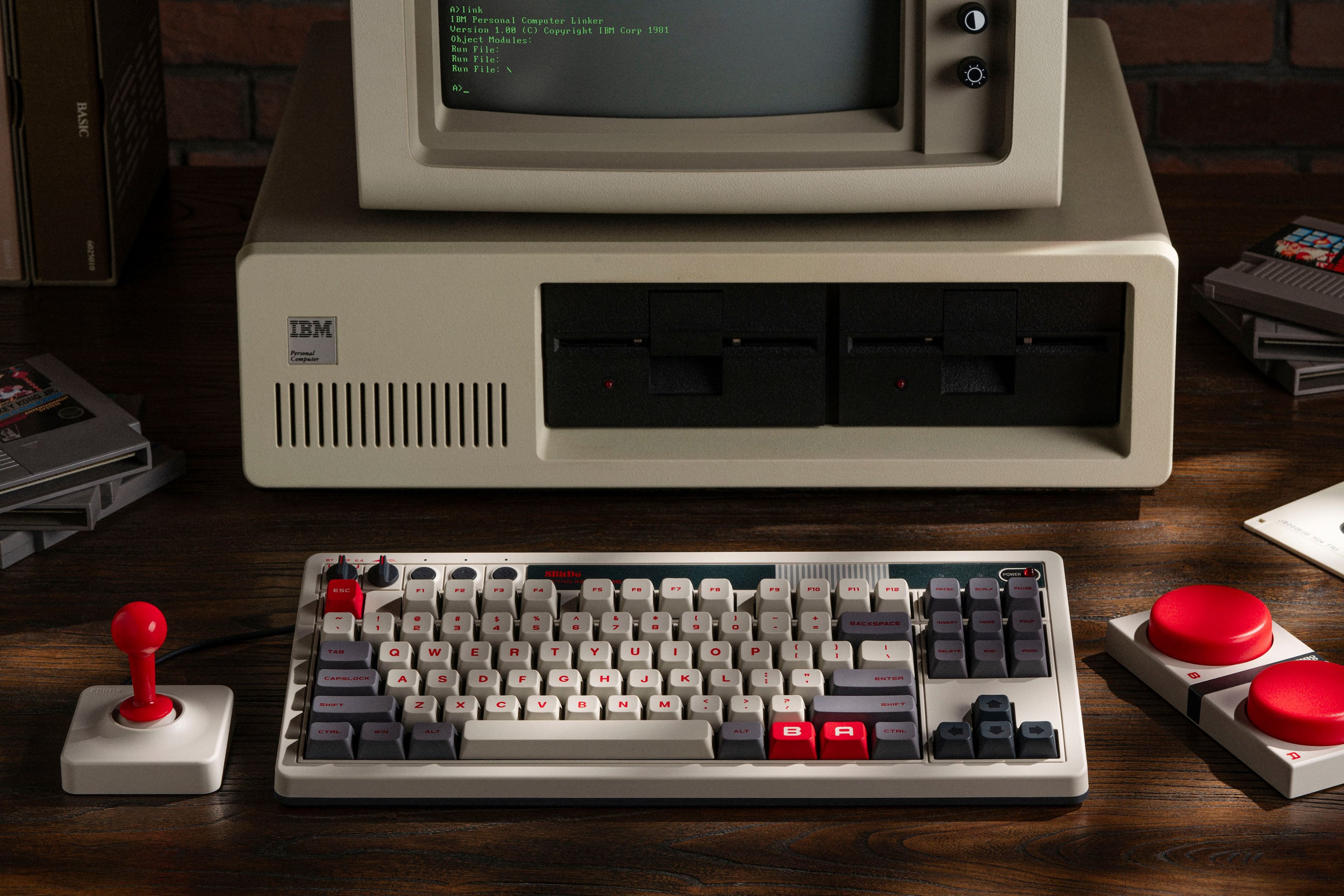 The image features a vintage IBM computer with a keyboard and a monitor. The computer is accompanied by a mouse, and there are several video game controllers scattered around the desk. The text on the image reads "IBM Personal Computer Lisker Version 1.88 Copyright 1981 IP Corp." This suggests that the computer is an older model, likely from the 1980s, and it may have been used for both work and entertainment purposes.