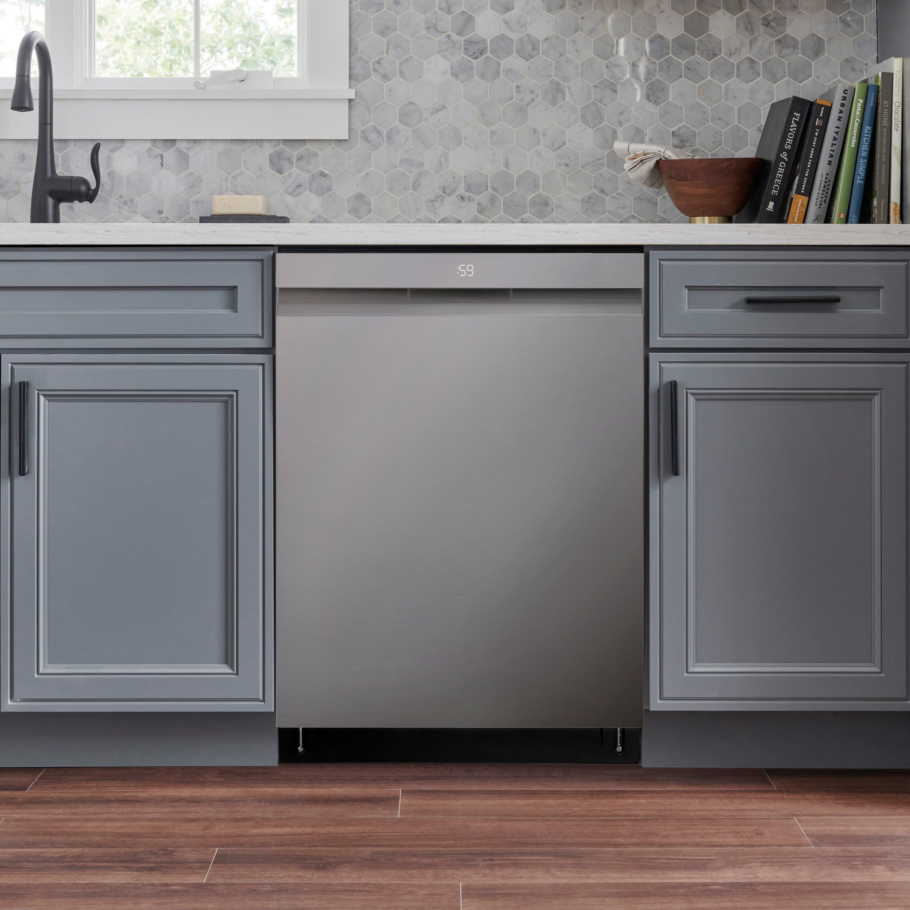 The image features a kitchen with a stainless steel dishwasher and a sink. The dishwasher is located under a counter, and there is a bowl on the counter. The kitchen also has a window, and a book is placed on the counter.