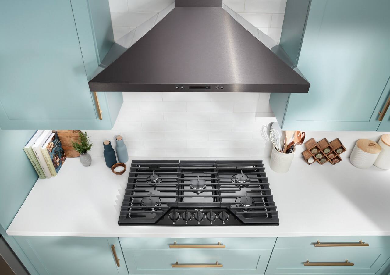 The image features a kitchen with a stove top oven, a microwave, and a sink. The stove top oven has a black cooking surface, and there is a large vent hood above it. The kitchen counter has various items on it, including a collection of books, a potted plant, and a vase. Additionally, there are several cups and bowls placed around the kitchen, and a knife is visible on the counter. The overall scene is clean and well-organized, with a focus on the stove and its surroundings.