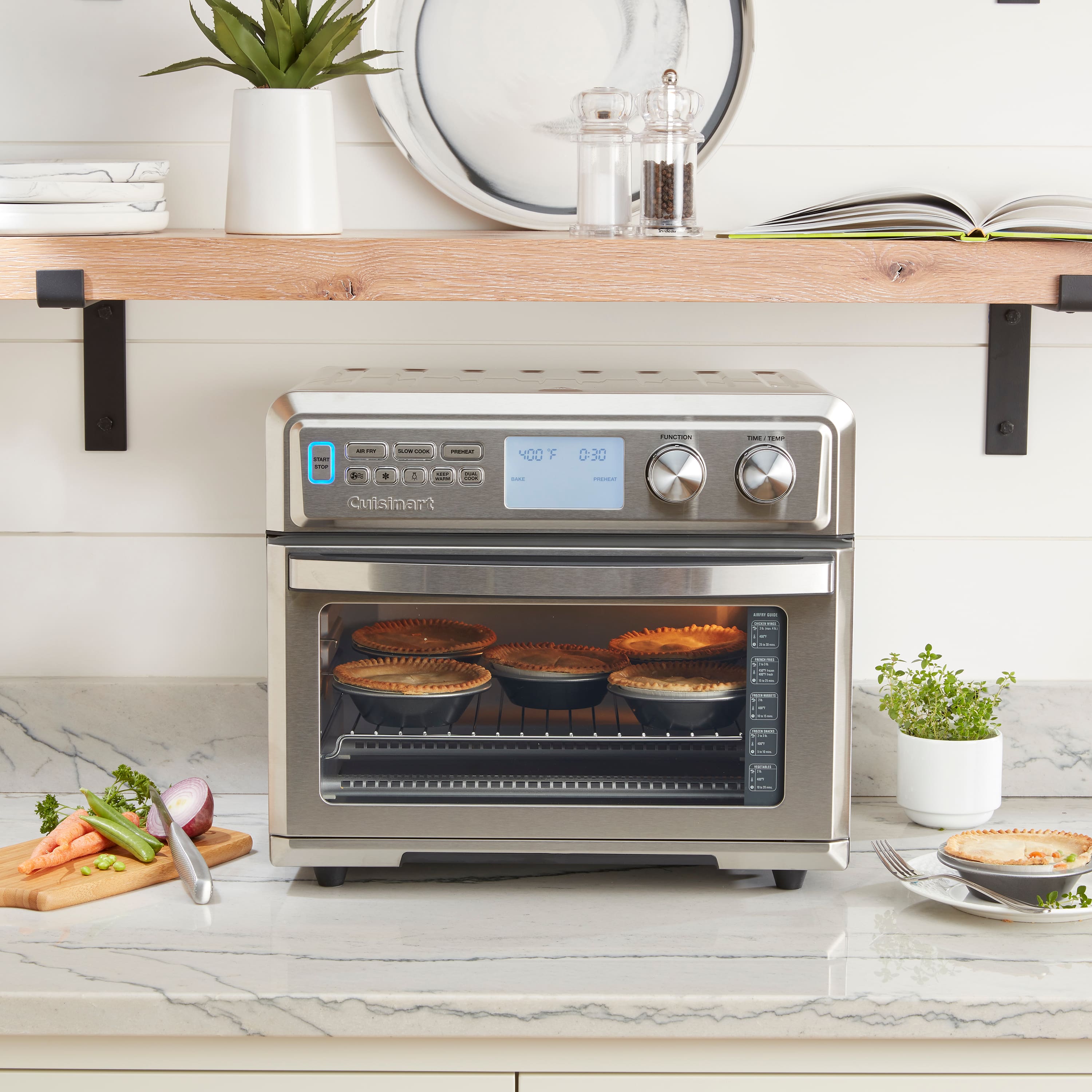 The image shows a silver oven with a blue display, which is currently set to 300 degrees. The oven is filled with a variety of food items, including pies and casseroles. There are several pies placed in different positions within the oven, and a casserole is also visible. The oven is situated on a countertop, and there are multiple books nearby, possibly cookbooks or other reading materials. Additionally, there is a potted plant and a fork placed on the countertop, adding to the overall ambiance of the kitchen scene.