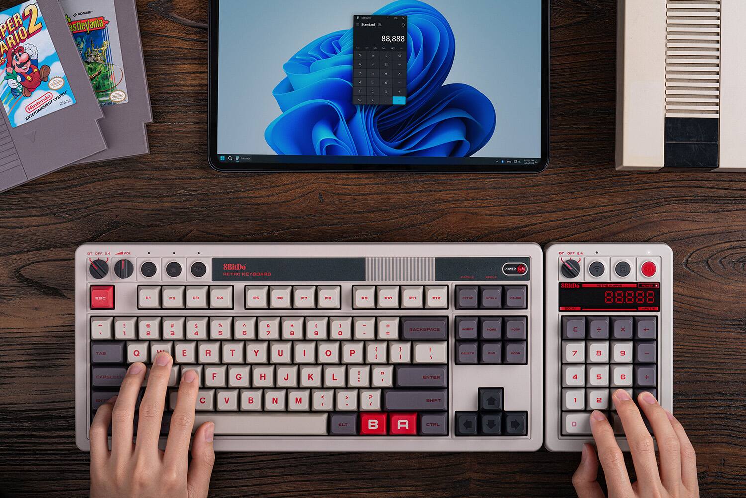 The image shows a person using a computer with two keyboards, one of which is a Nintendo keyboard. The person is typing on the keyboards, and there is a laptop screen in the background. The text on the image reads: "D MontaR PER 2 astleyania ARIO IS + Swidand 88,888 - 1 - - + . - - - 1 . SYSTEM ENTERTAINMENT Nintendo - 1 - . - 1 - e - i . A VIL n EFT . SBIDo RETIO RETMIAFIC 0 OAL ESC F1 F2 F. F4 FS FB F7 F F9 F10 .1 F10 PCE . - Nitiio - - - 88888 8 - - A I 1 . 2 a a 5 4 S 5 a N & 7 . U I g I O I + - BACESPALE N - FL C + x - TAB Q E R T Y U I O P 1 1 1 1 . . . - - 7 8 9 - CAPBLOCK F G H J K L - - 1 ENTER 4 5 6 + SI C V B N M C > ? I SUIFT 1 2 ALT B A CTAL O."