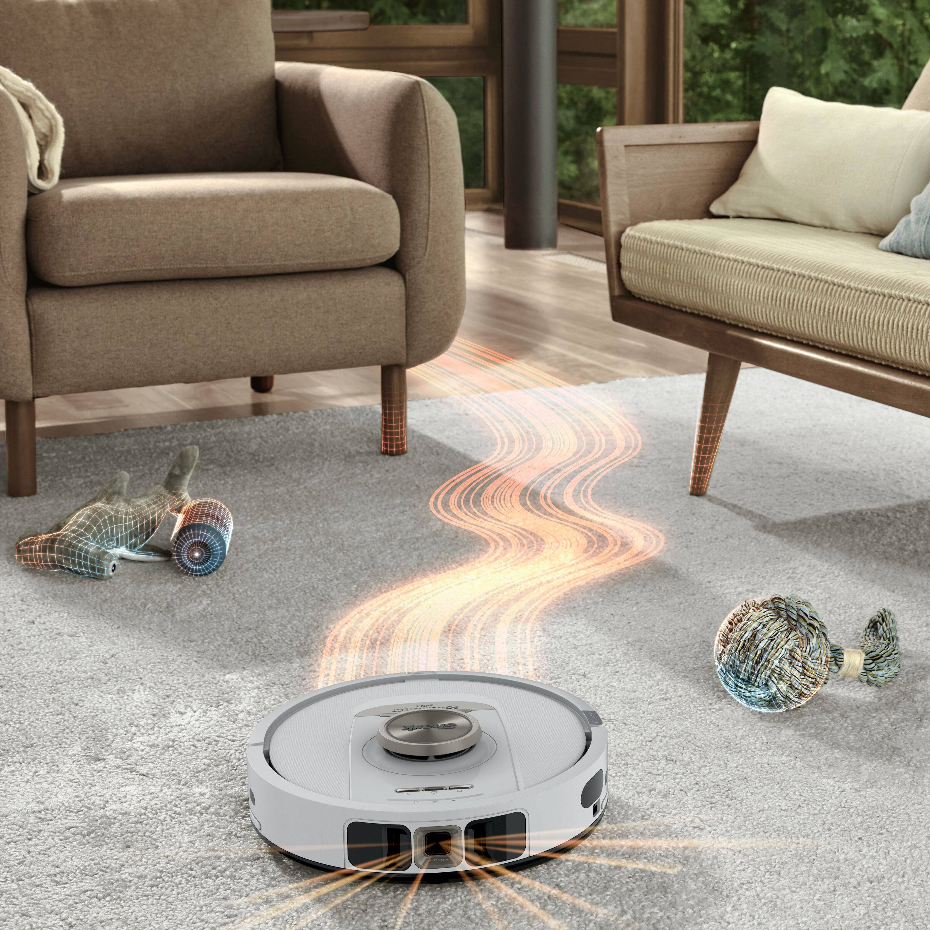 The image shows a robotic vacuum cleaner navigating a carpeted area in a living room. The vacuum cleaner is white with a circular design and has sensors and a central brush visible on its top. It is surrounded by animated orange lines indicating its path and mapping of the area. There are two small, spherical objects on the carpet, possibly toys or decorative items. The room features a brown armchair on the left and a beige sofa on the right, with a wooden floor and a green outdoor area visible in the background.