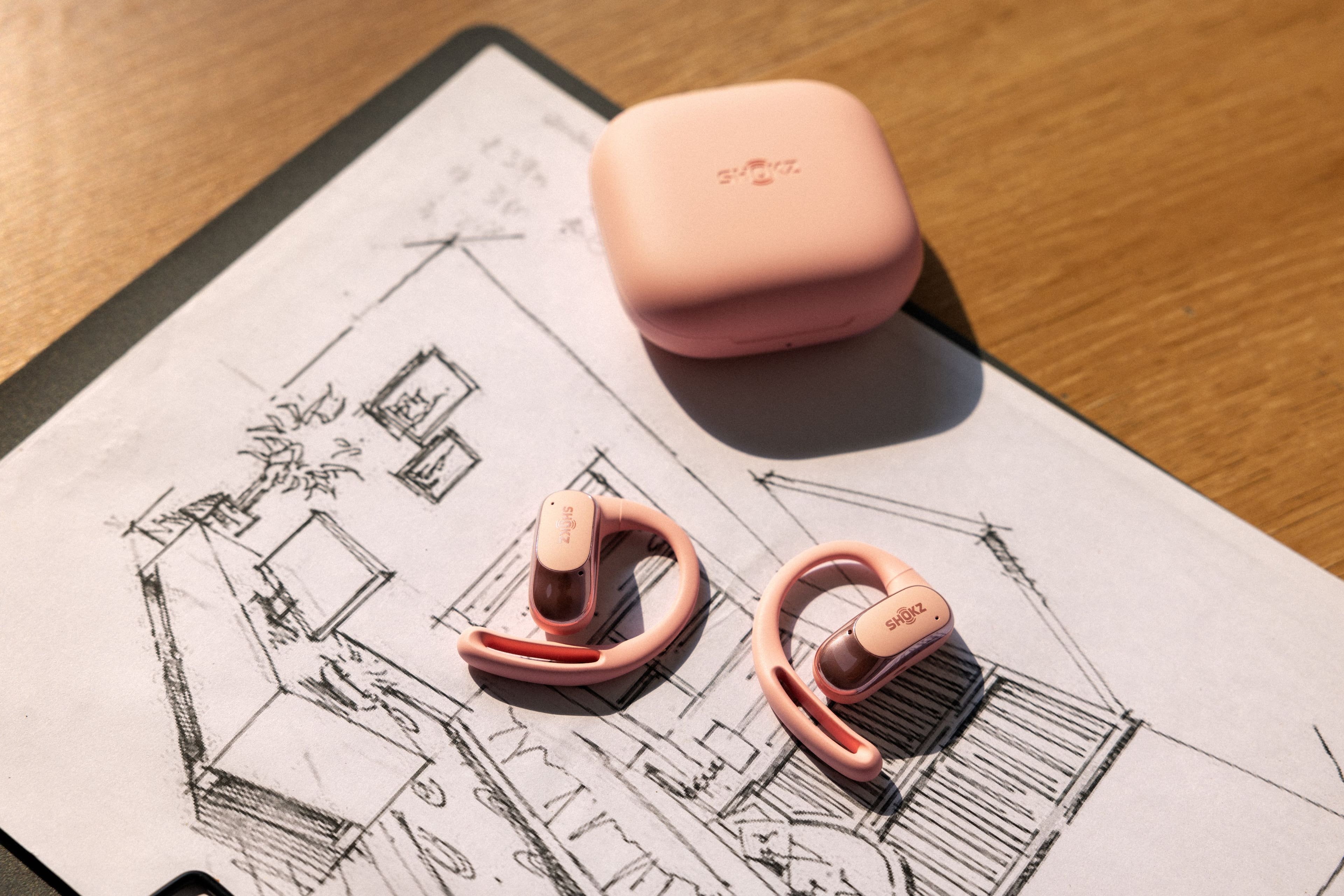 The image features a pair of pink earbuds sitting on top of a piece of paper. The earbuds are accompanied by a small box, which is likely the charging case for the earbuds. The box is placed next to the earbuds, and both are situated on a wooden table. The earbuds are designed to be worn in the ears, providing a comfortable and secure fit for the user.