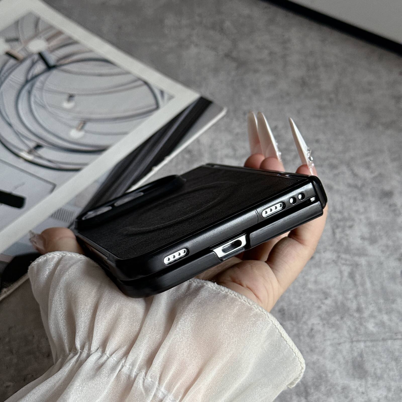 The image shows a person holding a black smartphone with a textured back cover. The phone has a camera module with three lenses and an LED flash. The person is also holding a pair of white nail extensions. In the background, there is a magazine or book with a circular design on the cover.