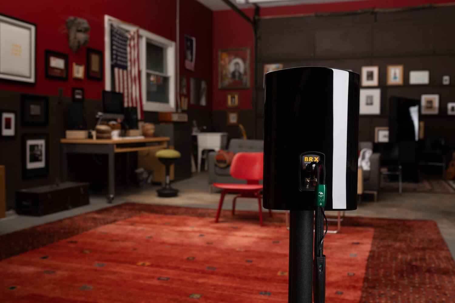 The image features a room with a red rug and a black and white striped machine. There is a chair in the room, and a desk with a computer on it. The room also contains a dining table and a TV. The walls are adorned with various pictures and a flag. The overall atmosphere of the room appears to be a mix of a living space and a workspace.