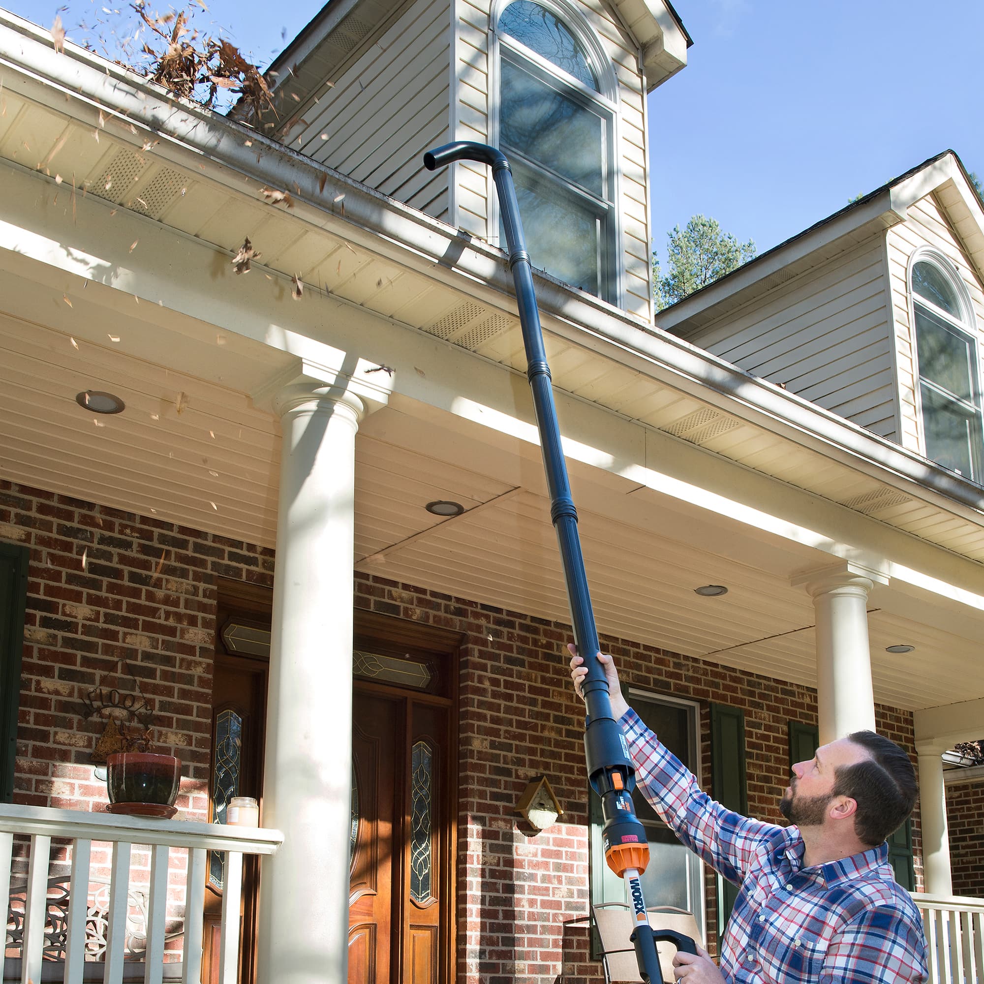 A man is standing outside of a brick house, holding a leaf blower. He is using the leaf blower to clean the leaves off the roof of the house. The man is wearing a plaid shirt and is focused on his task. The leaf blower is positioned above the house, and the man is actively operating it to remove the leaves from the roof.