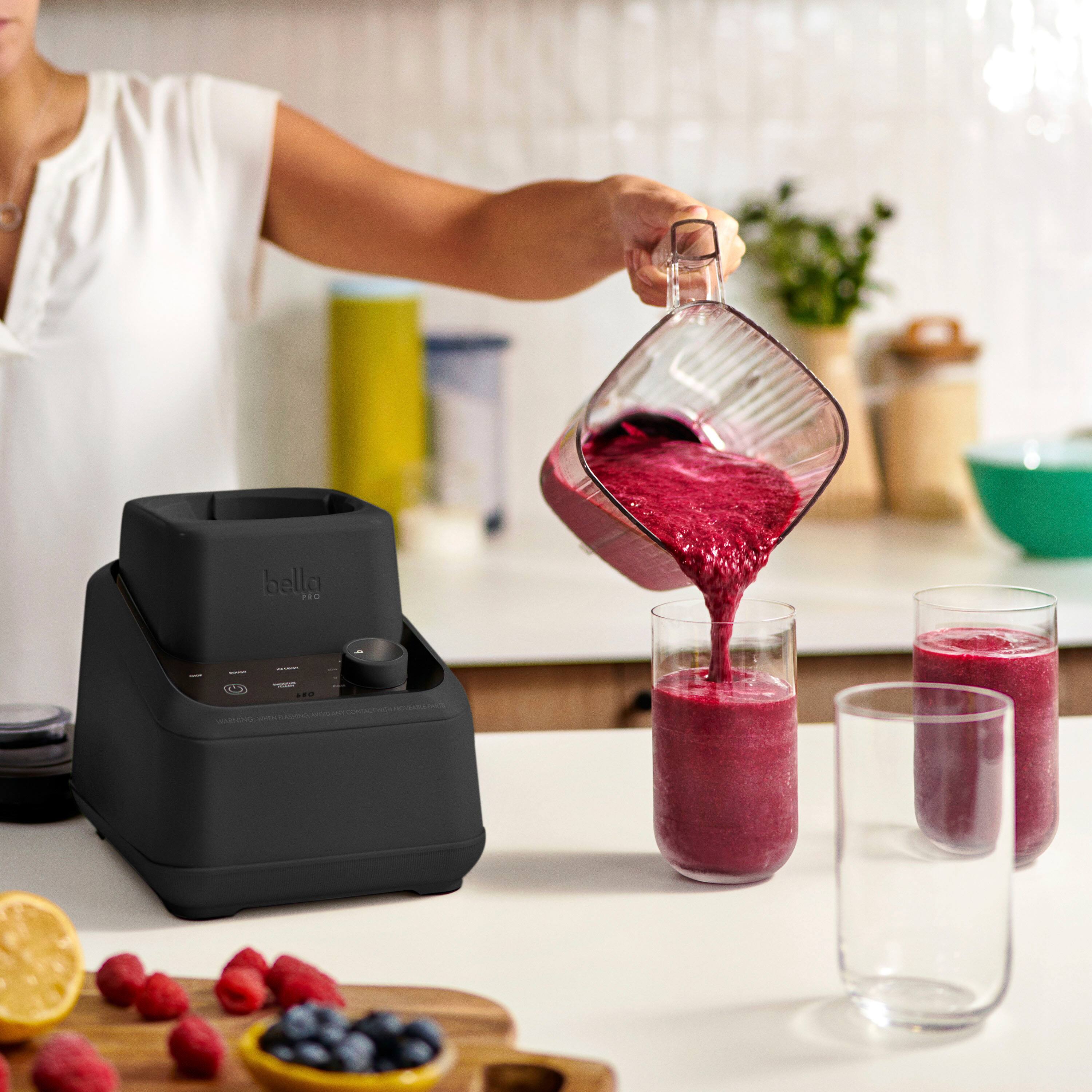 The image shows a woman pouring a drink into a glass using a Bella blender. The blender is placed on a counter, and there are two glasses nearby. The counter also has a bowl and a potted plant. The woman is pouring the drink into one of the glasses.