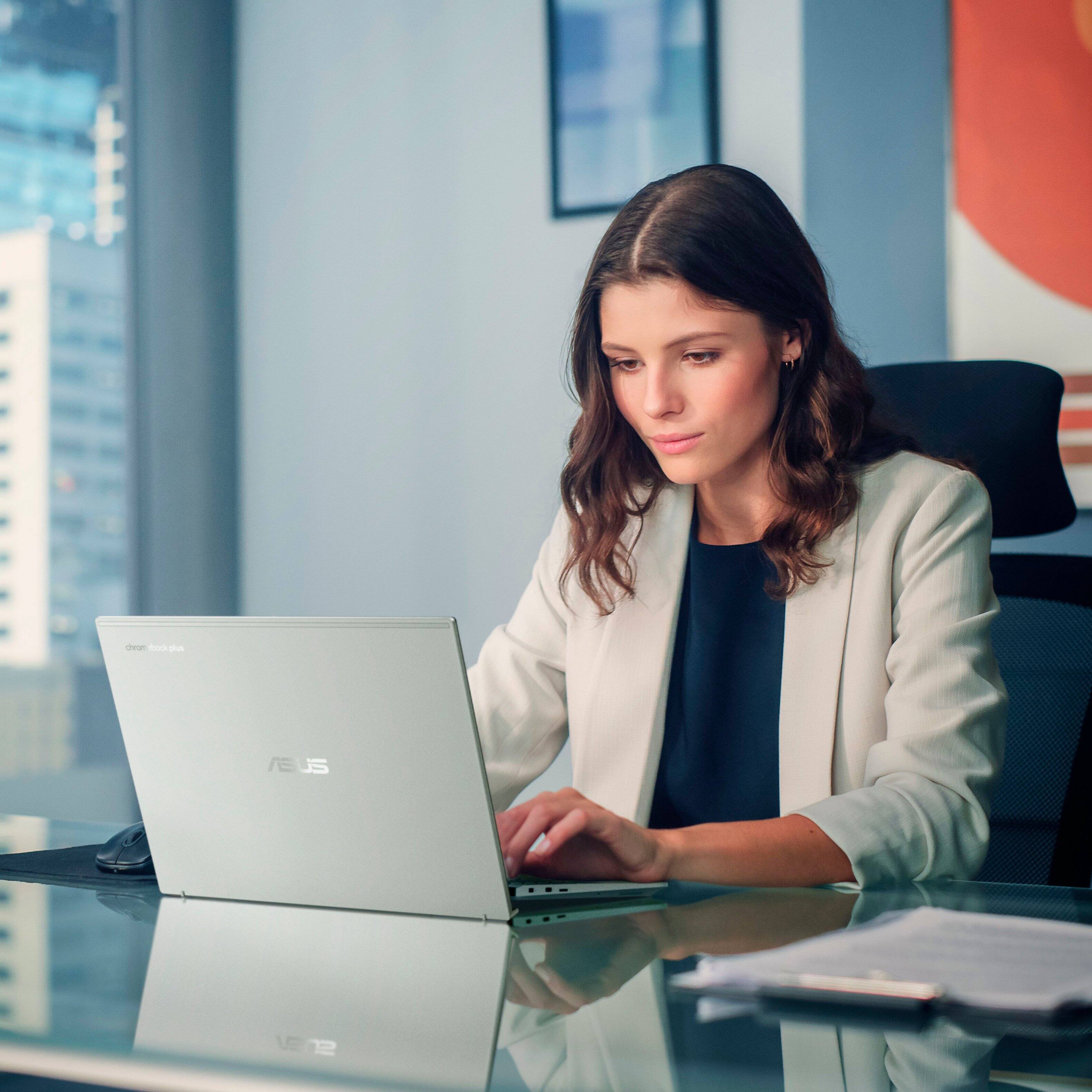 A woman is sitting at a desk, using a laptop. She is wearing a white jacket and a black shirt. The laptop is placed on a desk in front of her, and she is typing on the keyboard. The scene appears to be set in an office environment, with a chair visible behind the woman.