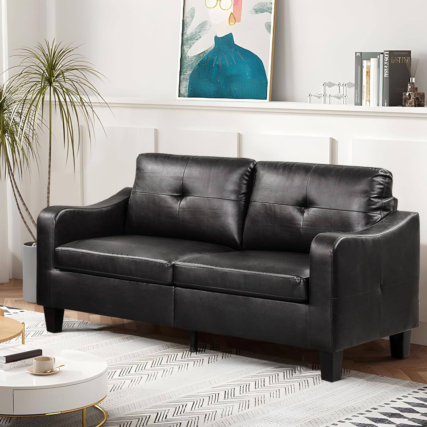 The image shows a black leather couch sitting in a living room. The couch is positioned in front of a white wall, and there is a painting hanging above it. The room also features a potted plant and a bookshelf with books on it. The couch is placed on a rug, and there is a coffee table nearby. The overall scene gives off a cozy and comfortable atmosphere.