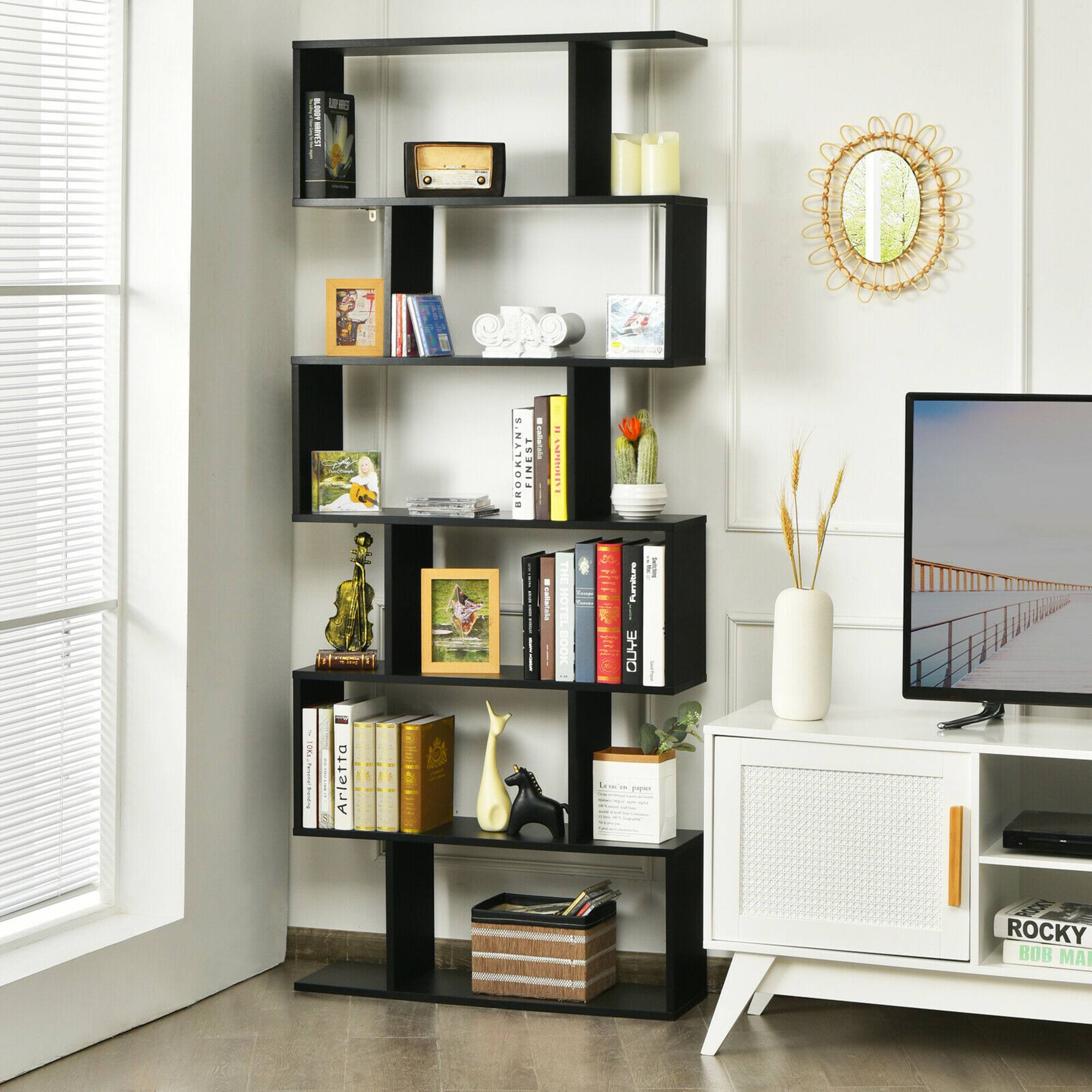 The image shows a living room with a television on a white stand. The room also features a bookshelf filled with books, a vase, and a mirror. The bookshelf is located near the television, and there is a window in the room. The television is turned on, and the room appears to be well-organized and comfortable.
