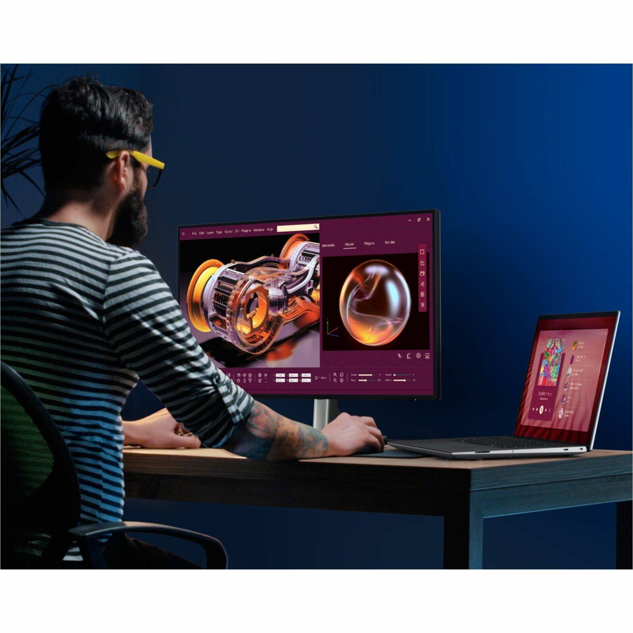 The man is sitting at a desk with a laptop and a large monitor. He is using a computer mouse to navigate the screen, which displays a 3D model of a car engine.