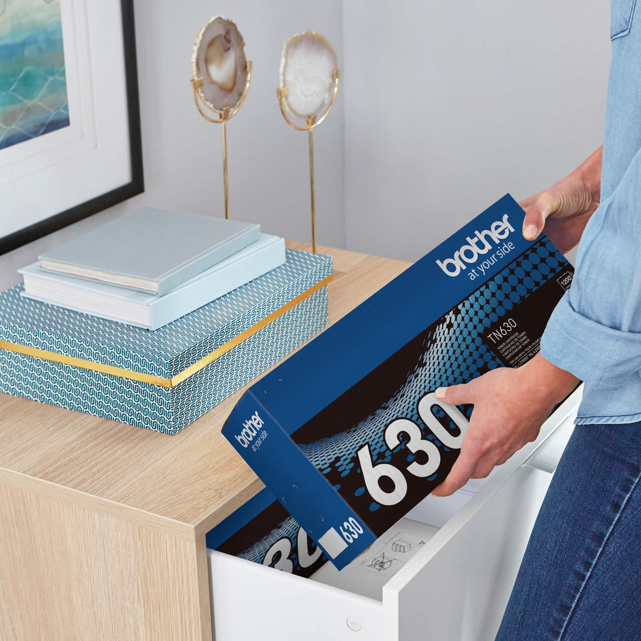 A person is holding a box with the text "brother at your side" on it. The box contains a printer, specifically the Brother TN630. The box is being opened, and the person is likely preparing to set up the printer.