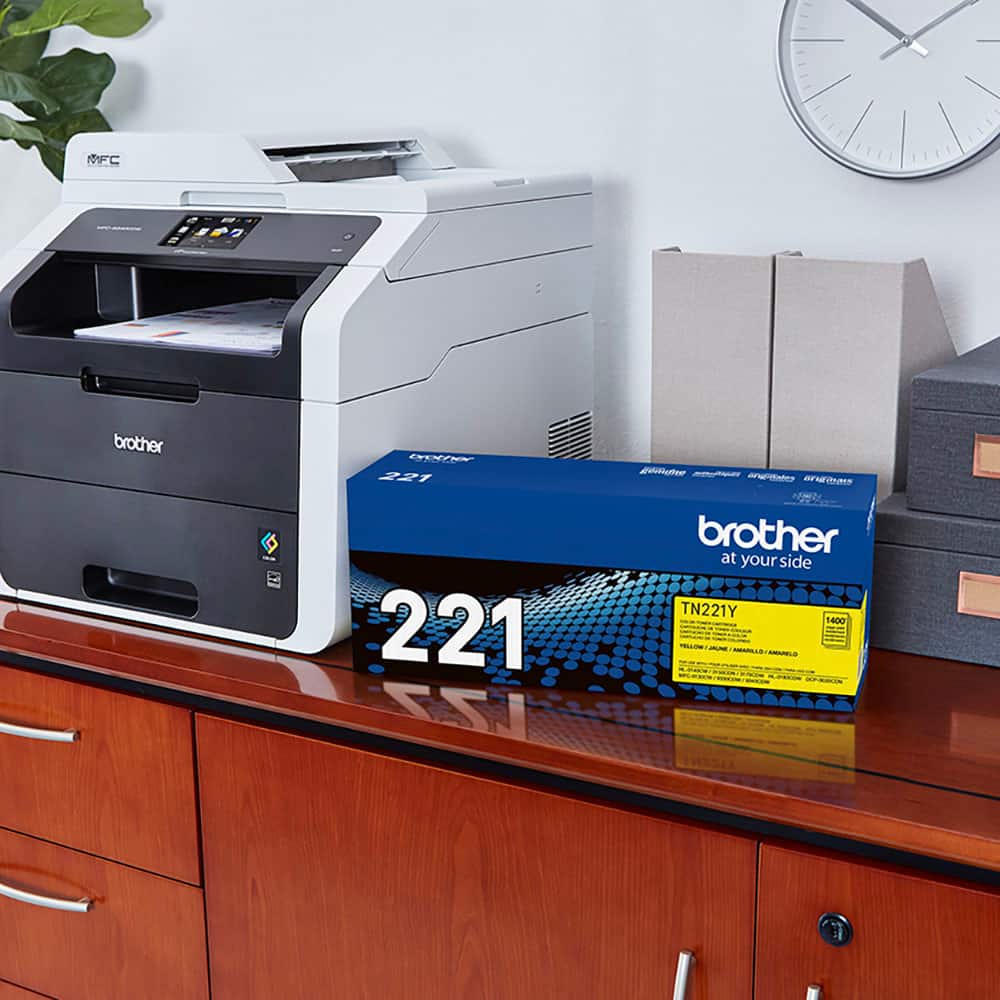 The image features a desk with a printer and a box of toner cartridges. The printer is a Brother MFC-221, and the toner cartridges are labeled as Brother at your side TN221Y. The box of toner cartridges is placed next to the printer, and there is a clock on the wall above the desk.