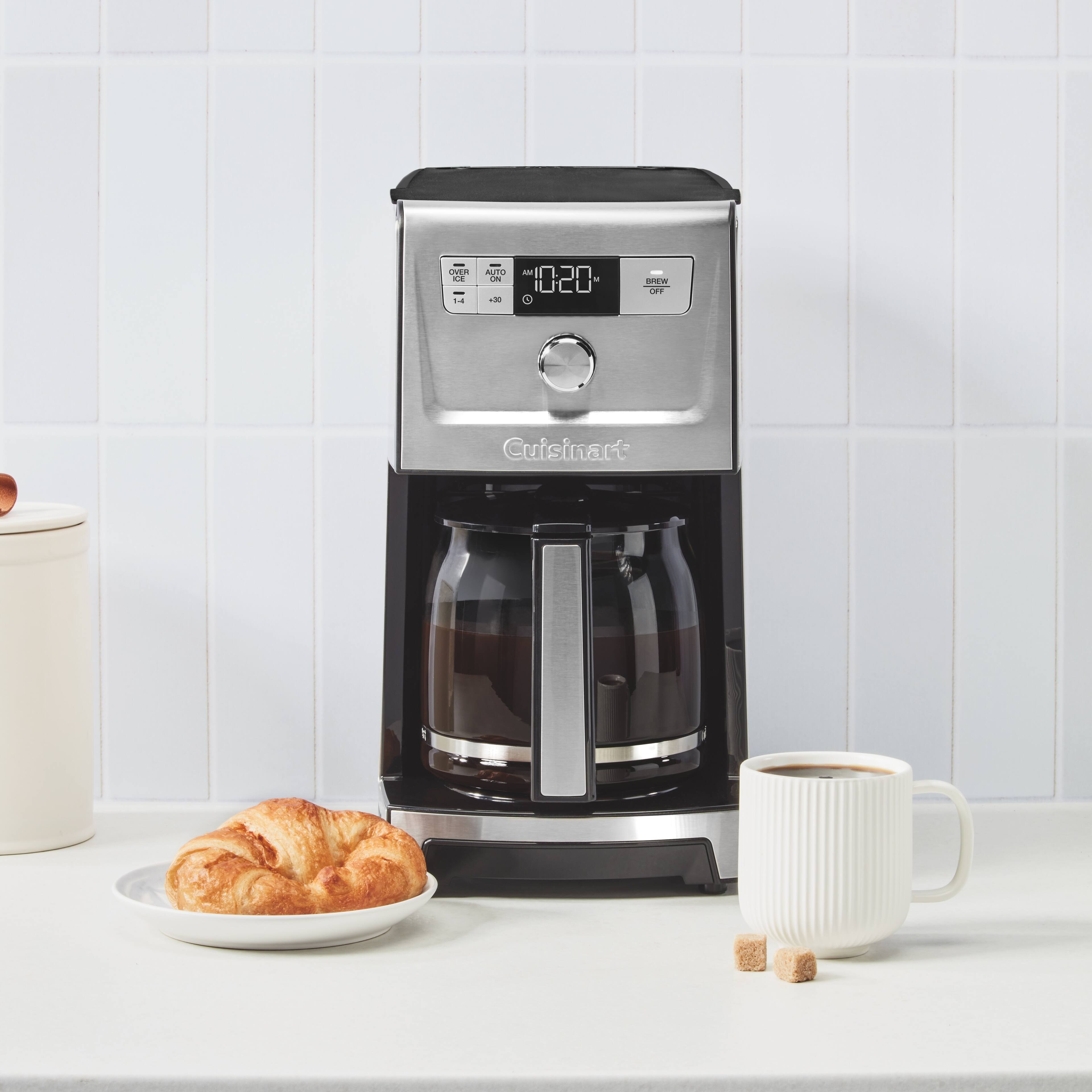 The image shows a Cuisinart coffee maker sitting on a counter next to a plate of croissants. The coffee maker is a silver and black appliance, and it is accompanied by a cup of coffee. The scene suggests a cozy morning or afternoon, with a delicious breakfast or snack being enjoyed alongside a warm beverage.