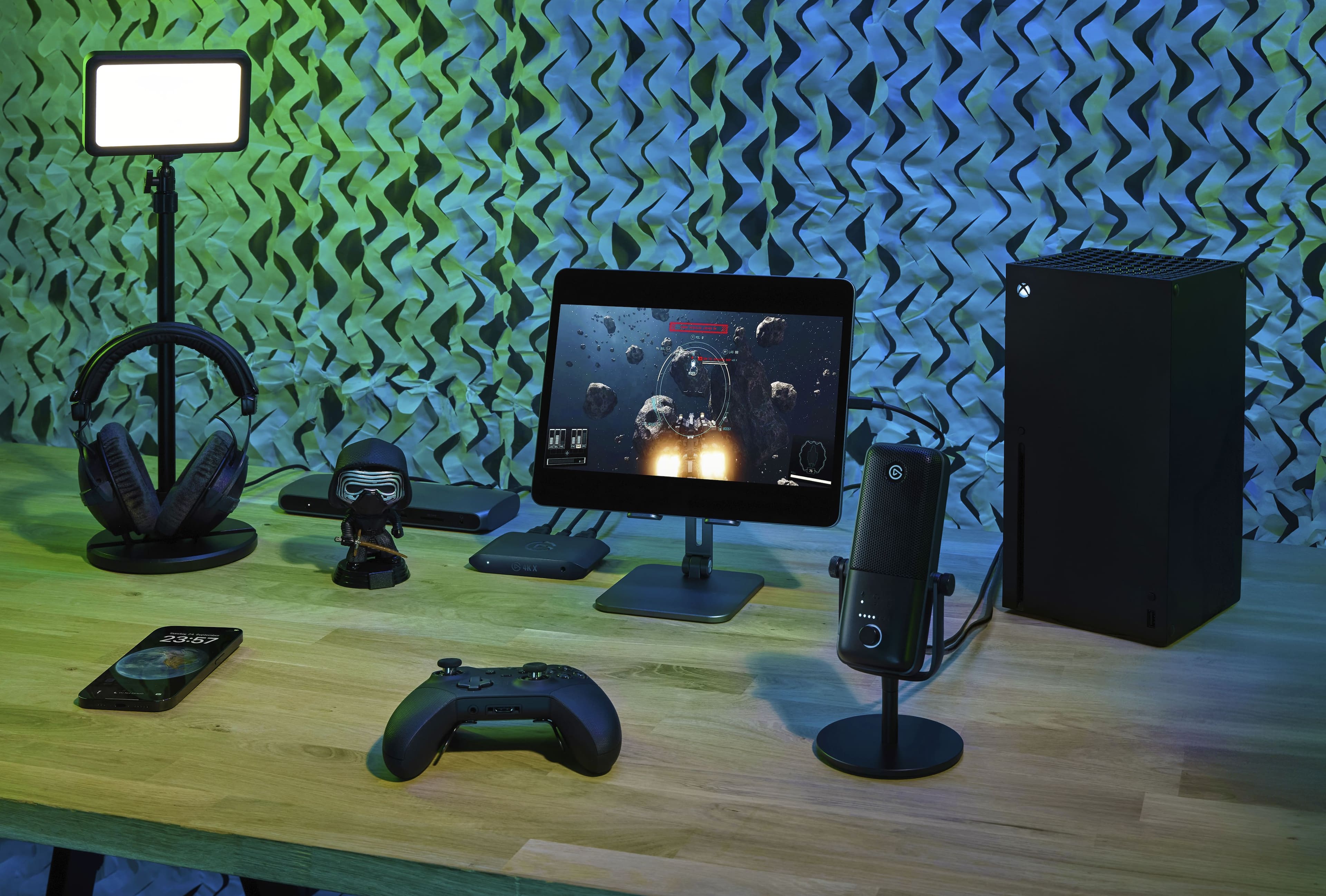 The image features a wooden table with various electronic devices on it. There is a laptop, a cell phone, a microphone, and a headset. The table also has a monitor, a keyboard, and a mouse. The scene appears to be set in a room with a green wallpaper.