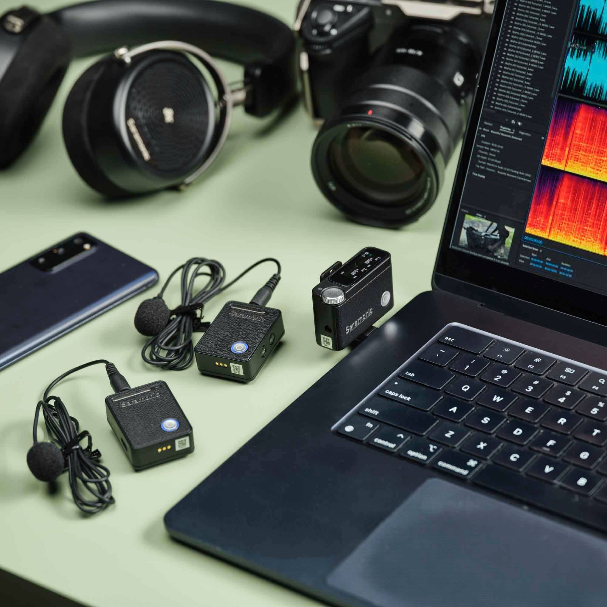 The image features a laptop computer sitting on a green surface, accompanied by various electronic devices. There are two cell phones, one on the left side of the laptop and the other on the right side. Additionally, there are two microphones, one placed near the laptop and the other on the left side. A camera is also present on the right side of the laptop. The laptop is open, and the arrangement of the devices suggests that they are being used for a project or recording.