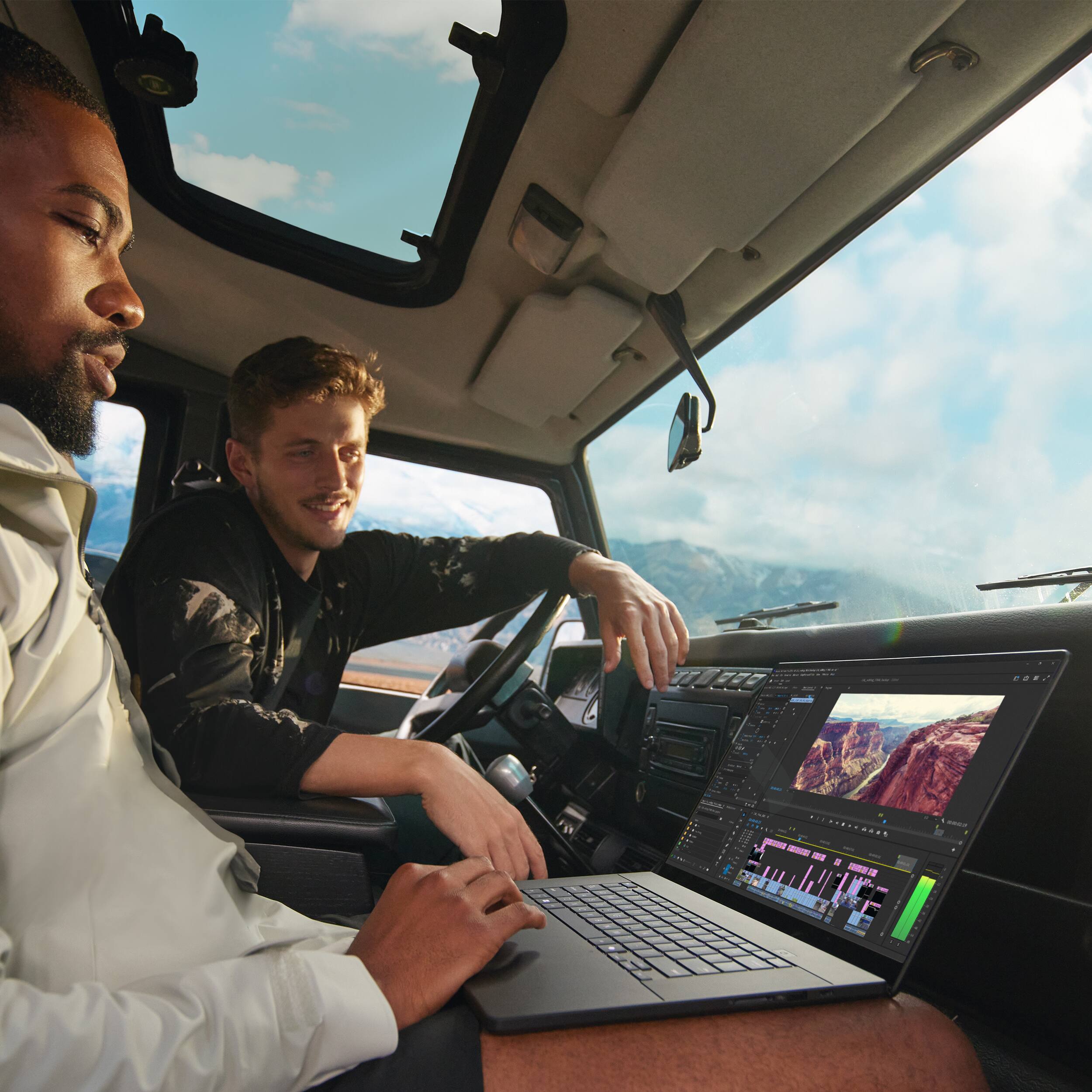 The image shows two men sitting in a car, with one of them using a laptop. The man using the laptop is wearing a white shirt and black shorts, while the other man is wearing a black jacket. The car appears to be a truck, and they are both smiling and enjoying their time together. The laptop is placed on the man's lap, and he seems to be focused on his work or activity. The scene captures a moment of relaxation and bonding between the two men.