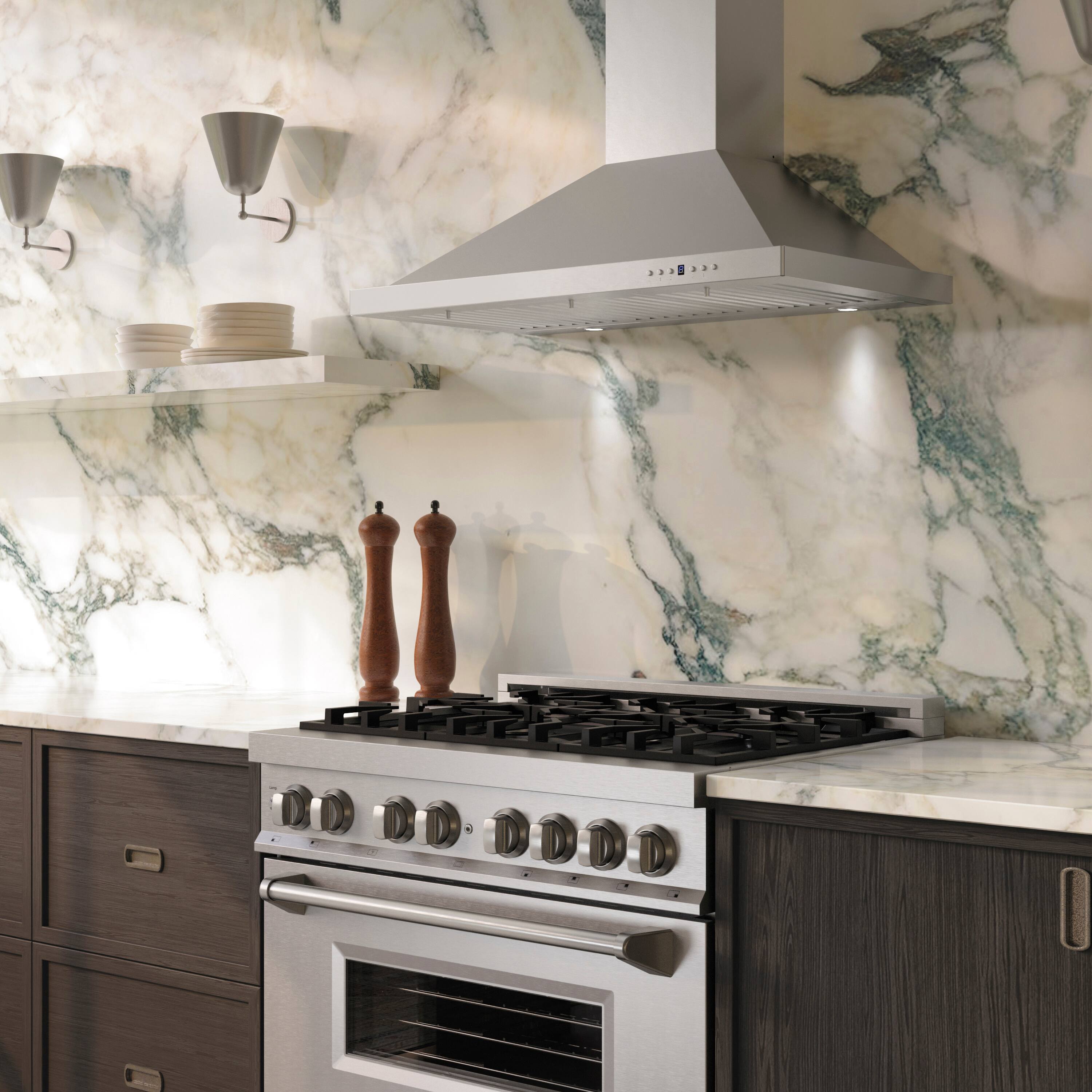 The image shows a modern kitchen with a stainless steel gas stove and oven. The backsplash and countertop are made of white marble with green veining. Above the stove, there is a stainless steel range hood. On the countertop, there are two wooden pepper mills. The cabinets are dark wood with metallic handles. There are also some plates stacked on a shelf above the countertop.