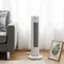 The image features a white portable air conditioner placed on a wooden floor near a window. The air conditioner is situated in front of a couch, and there is a picture of a banana leaf nearby. The scene appears to be set in a living room, with the air conditioner providing cooling for the space.