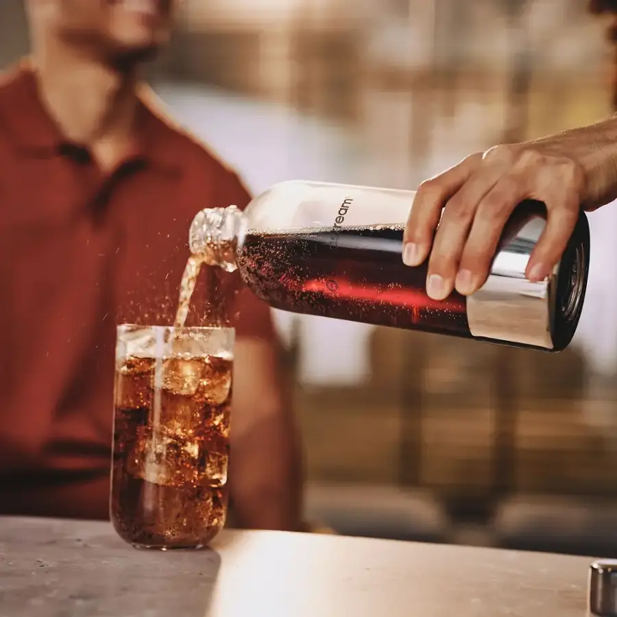 The image shows a man pouring a dark beverage, likely a soda, into a glass. The man is standing near a table, and the glass is placed on the table. The scene suggests that the man is enjoying a drink, possibly in a social setting or at home.