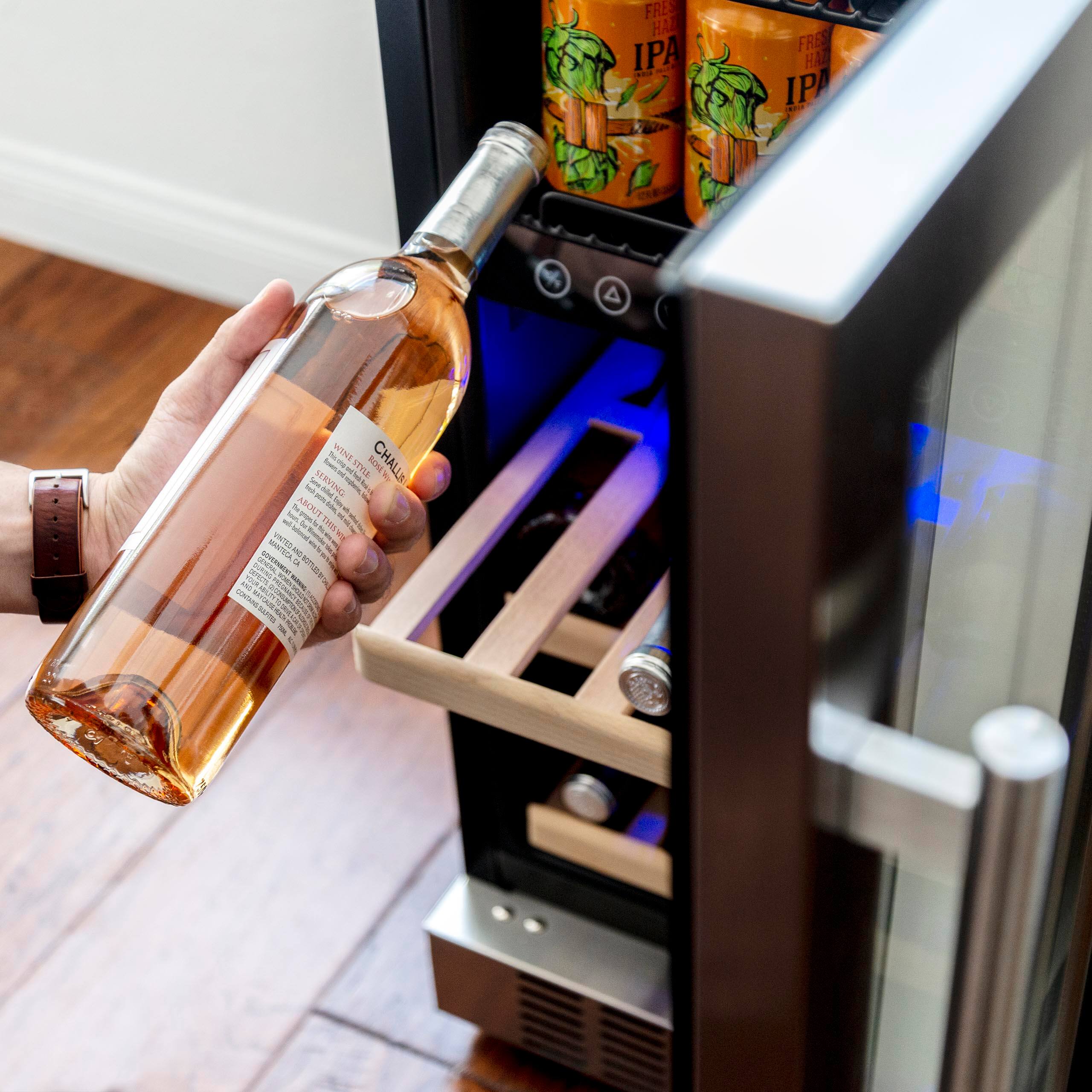 A person is holding a bottle of wine in front of a wine refrigerator. The wine refrigerator has a blue light inside and is filled with wine bottles. The person is likely selecting a wine to enjoy or store in the refrigerator.