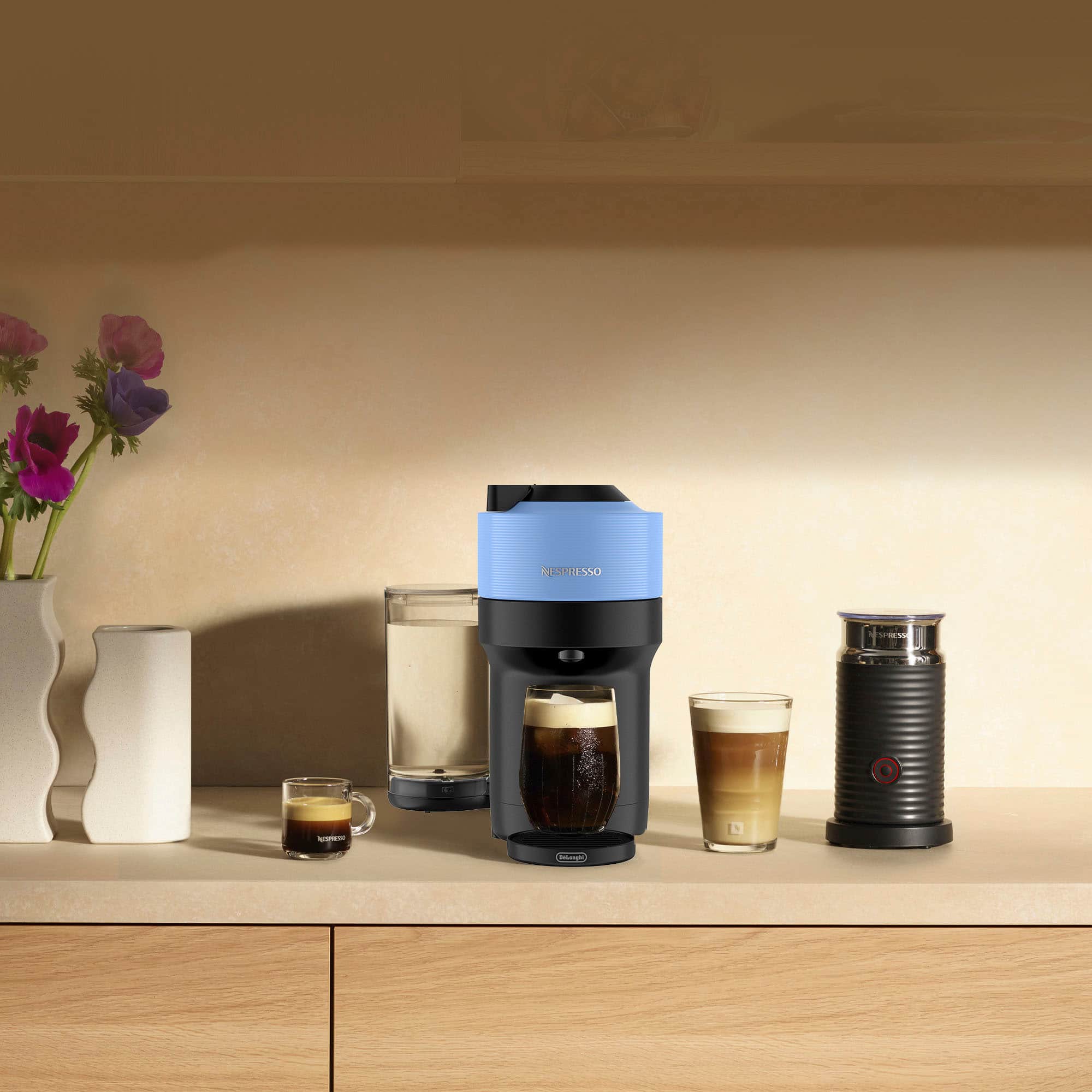 The image shows a counter with a coffee maker and a few cups. There are two cups of coffee, one on the left side and the other on the right side of the counter. Additionally, there are two glasses of beer, one near the center and the other towards the right side of the counter. A vase with flowers is also present on the left side of the counter. The coffee maker is positioned in the middle of the counter, and a bottle can be seen on the right side.