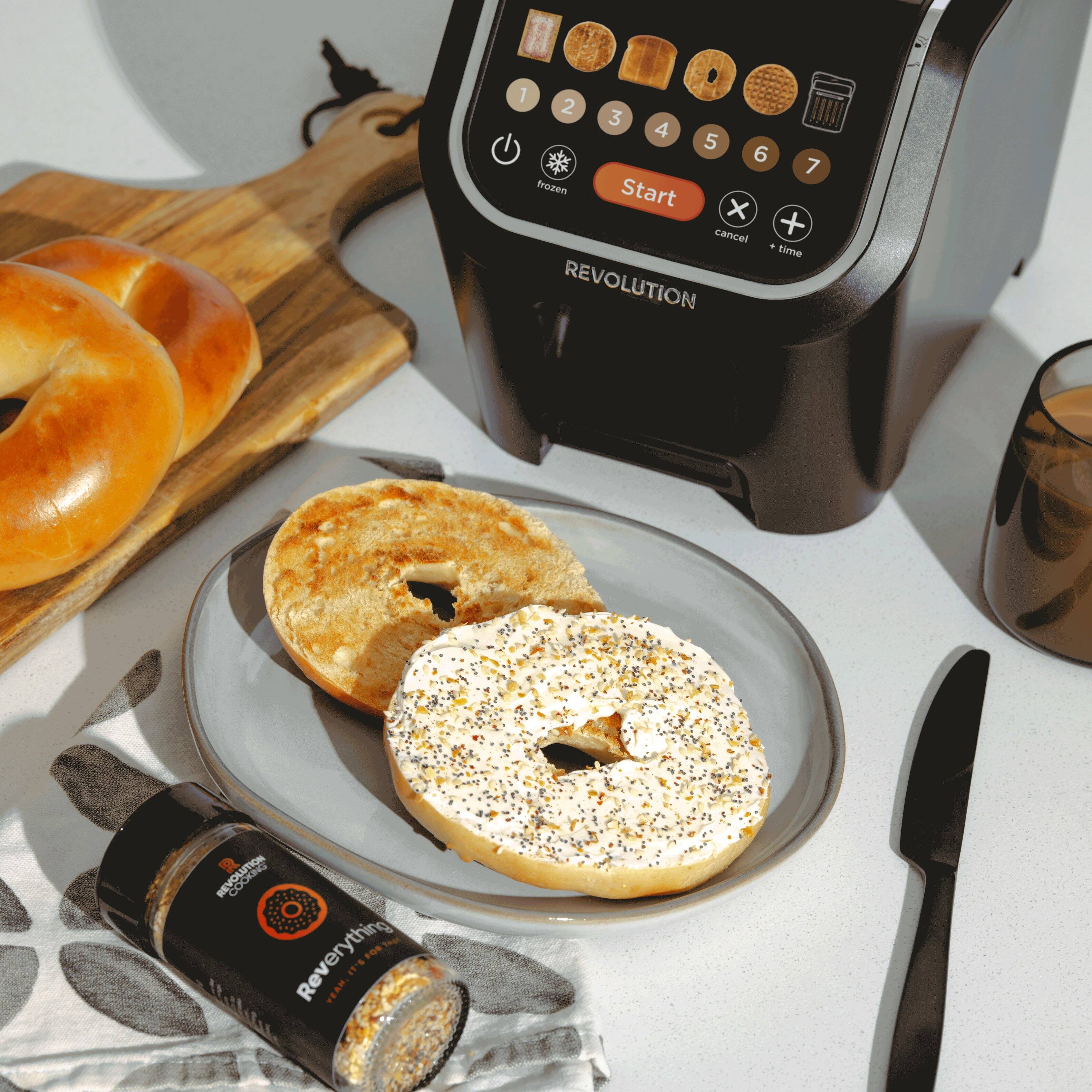 The image features a table with a bagel, a bagel sandwich, and a plate of bagels. There is also a toaster and a coffee cup on the table. The toaster is a Revolution toaster, and it is placed next to the bagels. The table setting includes a knife and a cup, suggesting that the meal is ready to be enjoyed.