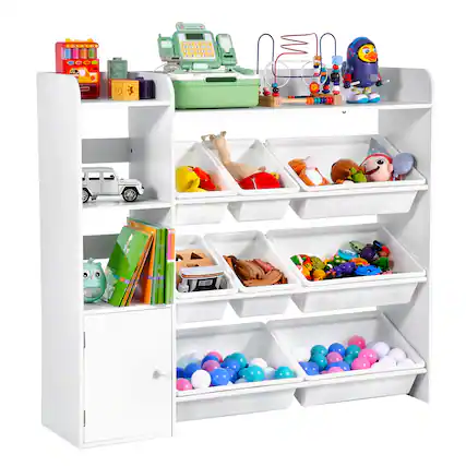 The image shows a white shelving unit filled with various toys and items. On the top shelf, there is a red toy cash register, a green toy cash register, and a blue and white bird-shaped toy. Below that, there are compartments with different toys, including a silver toy car, a yellow and red toy, a brown stuffed animal, and a green and white toy. The middle shelf has a green book with an owl on the cover, a small toy, and a compartment with colorful balls. The bottom shelf has two compartments filled with blue, pink, and white balls.