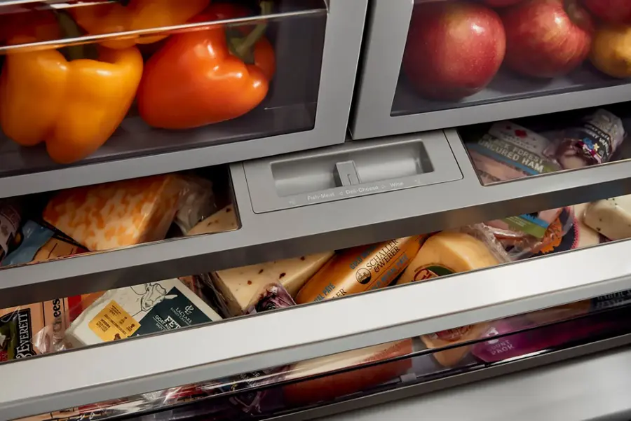 The image shows an open refrigerator with various food items inside. There are several apples placed in the upper part of the refrigerator, and a drawer filled with cheese and meat. The refrigerator also contains a few bottles, possibly containing wine or other beverages. The refrigerator is well-stocked with a variety of food items, making it a convenient and accessible source of nourishment for the household.