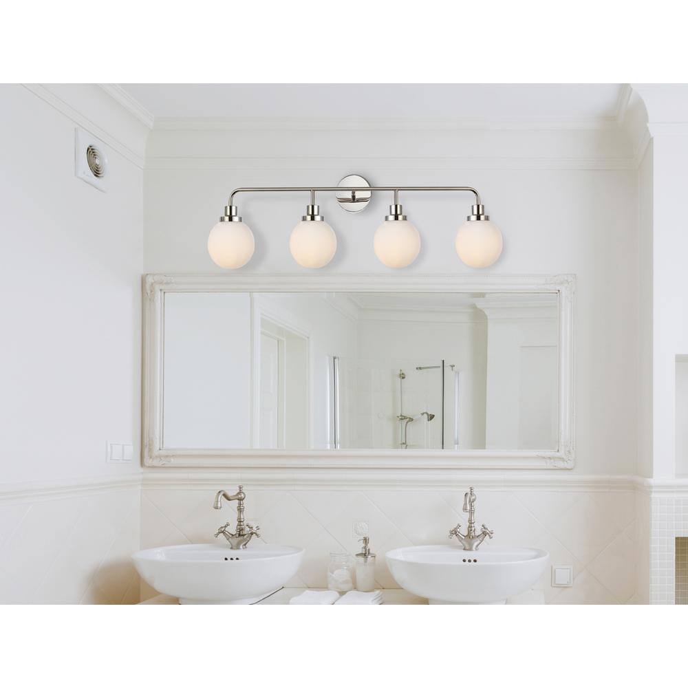 The image shows a bathroom with a large mirror above two white vessel sinks. The sinks have silver faucets, and there is a light fixture with four bulbs above the mirror. The walls are white, and there is a towel and some toiletries on the counter.