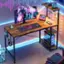 The image features a computer desk with a keyboard, mouse, and monitor. There is a laptop on the desk as well. The desk is situated in a room with a chair and a TV. The room also has a shelf with a cell phone and a bottle on it. The desk is set up in a dark room, and there is a keyboard with a pink light on it.