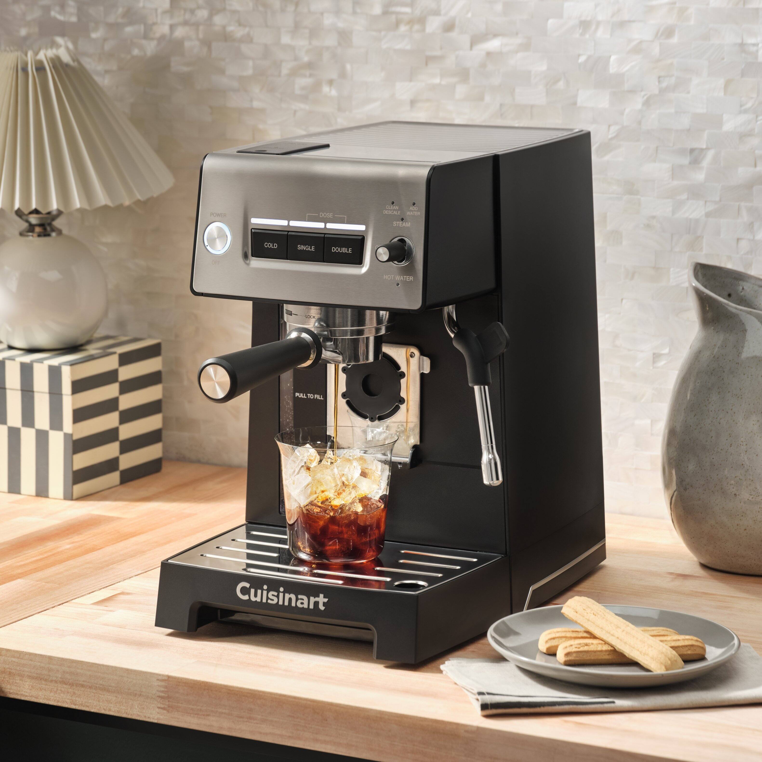 Cuisinart is a brand that manufactures kitchen appliances, including coffee makers and espresso machines. In the image, there is a Cuisinart espresso machine on a table, with a cup of coffee being made. The machine is designed to make both hot and cold coffee, and it can also steam milk for use in various coffee drinks. The espresso machine is placed on a wooden table, and there is a plate of cookies nearby, adding to the cozy and inviting atmosphere of the scene.