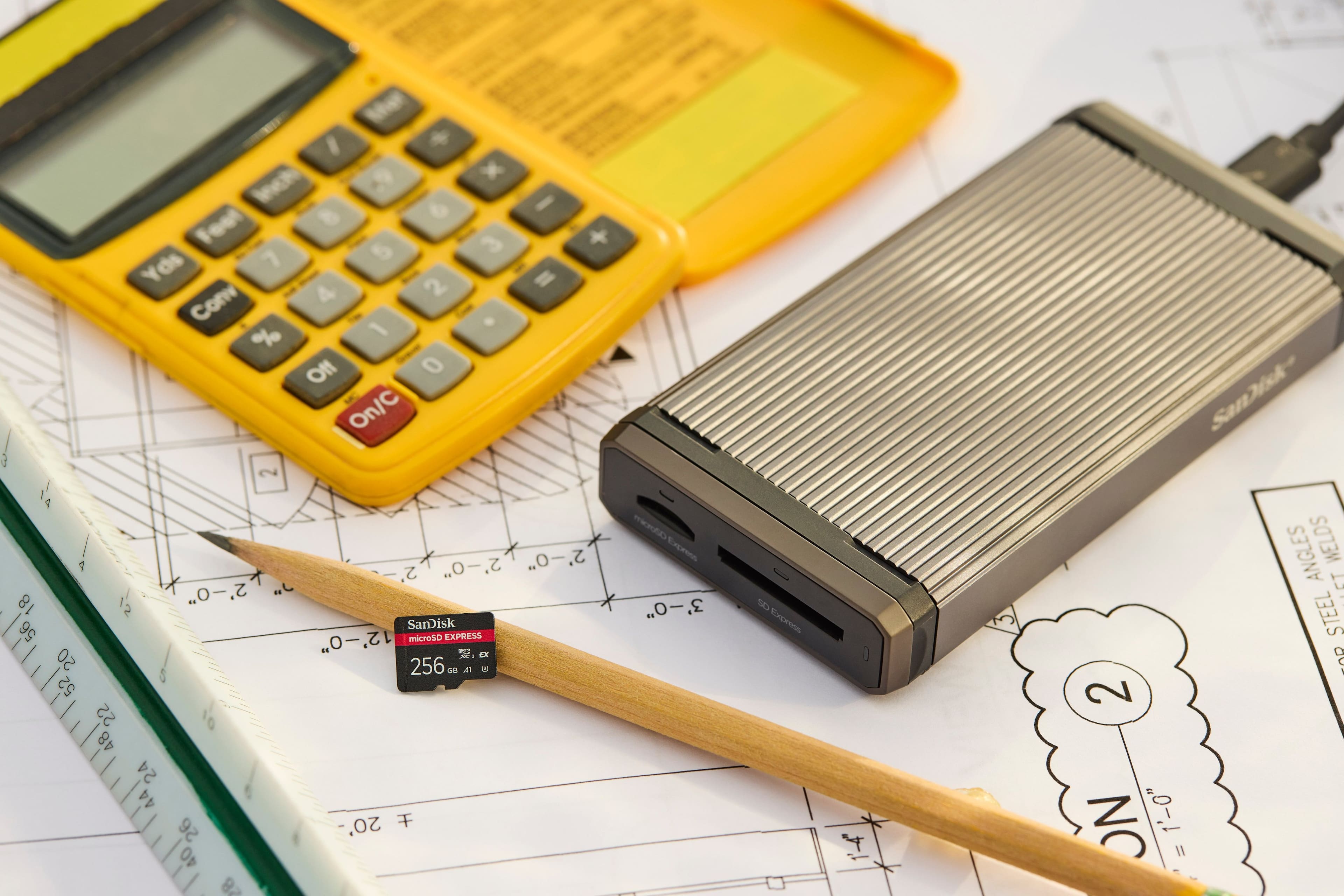 The image shows a calculator, a microSD card, and a pencil on top of a piece of paper. The text on the image reads: "x e I Foot 3 + Yds 7 2 I Con 4 % 1 Of 0 On/C a 2 1 airoen 2'-0" .O 2' 2'-0" .O 2' 2'-0" .O 2' 18 12 3'-0" 3 SanDisk 56 2'-0" | 2' microSD EXPRESS O m aX I 20 256 ca a 52 22 10 48 24 44 20' 26 40 e 28 S0 Express 4 Sanisk ANGLES WELDS rT STEEL : 2 NO 1'-0" II."