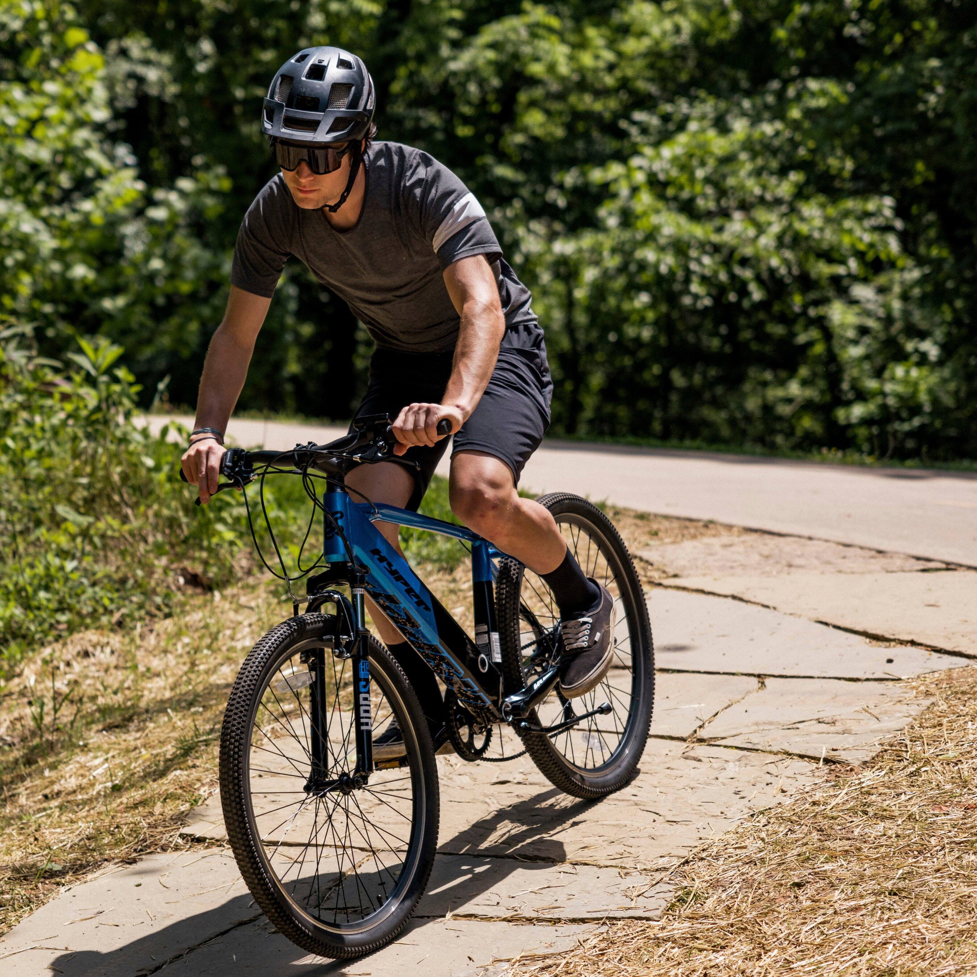 The man is riding a bicycle on a path through the woods. He is wearing a helmet for safety and is focused on the road ahead. The bicycle has a blue frame and is equipped with a basket for carrying items. The man is also wearing a gray shirt and black shorts while riding.