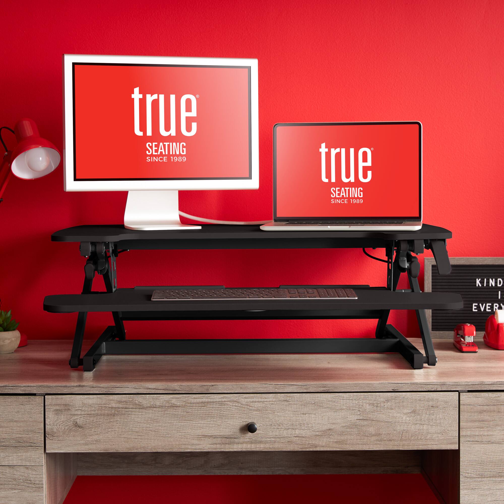 The image displays a desk with two laptops on it. The desk is red, and the laptops are placed on a black stand. The laptops are positioned in front of a monitor, creating a workstation setup. The desk also has a keyboard and a mouse, further enhancing the workspace.