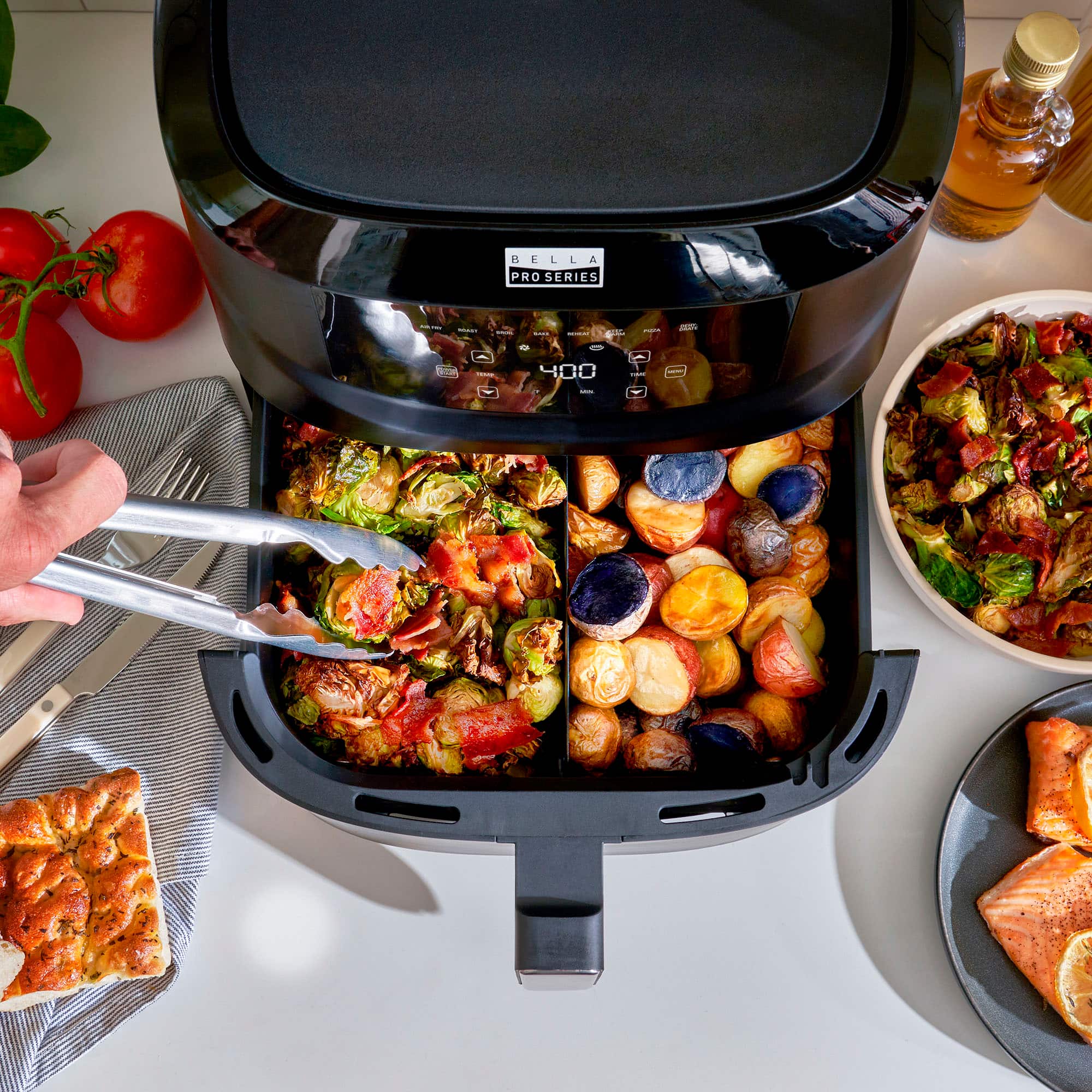 The image features a Bella Pro Series 1 toaster oven filled with various vegetables, including broccoli, tomatoes, and potatoes. The toaster oven is being used to cook the vegetables, and a person is in the process of taking a piece of broccoli out of it. The scene also includes a fork and a knife on the left side of the toaster oven, and a bowl of salad on the right side.