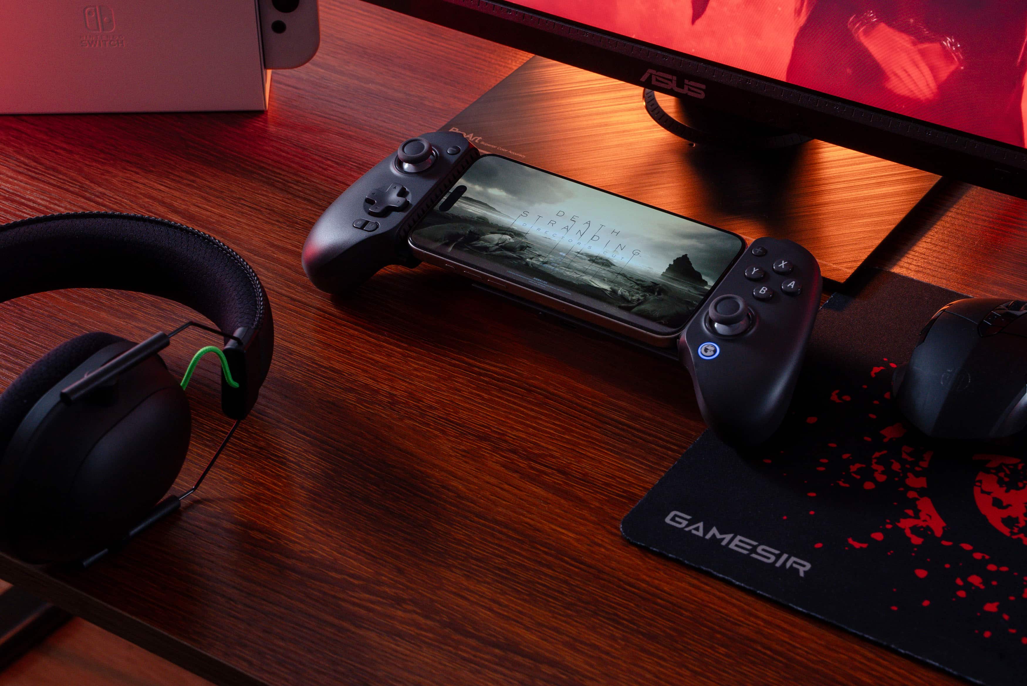 The image shows a wooden desk with a cell phone, two game controllers, and a pair of headphones. The cell phone is placed on top of the controllers, and the headphones are positioned on the left side of the desk. The scene suggests that someone is preparing to play a video game using the controllers and headphones.
